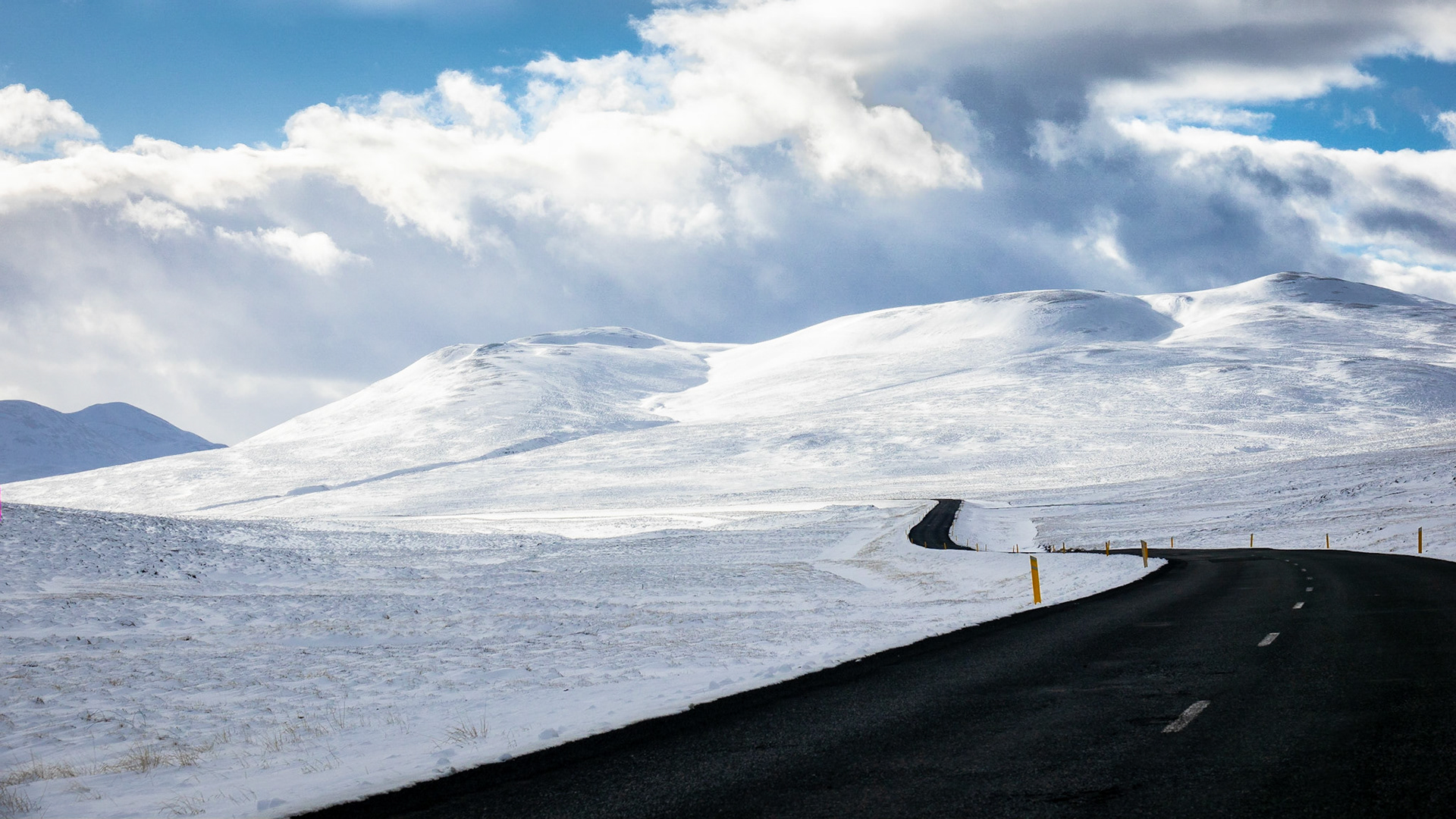 The winding black-topped asphalt highway of Route 1 - Iceland's Ring Road - weaves through a snowy landscape of fields and mountains under a blue sky with billowy but scattered clouds.