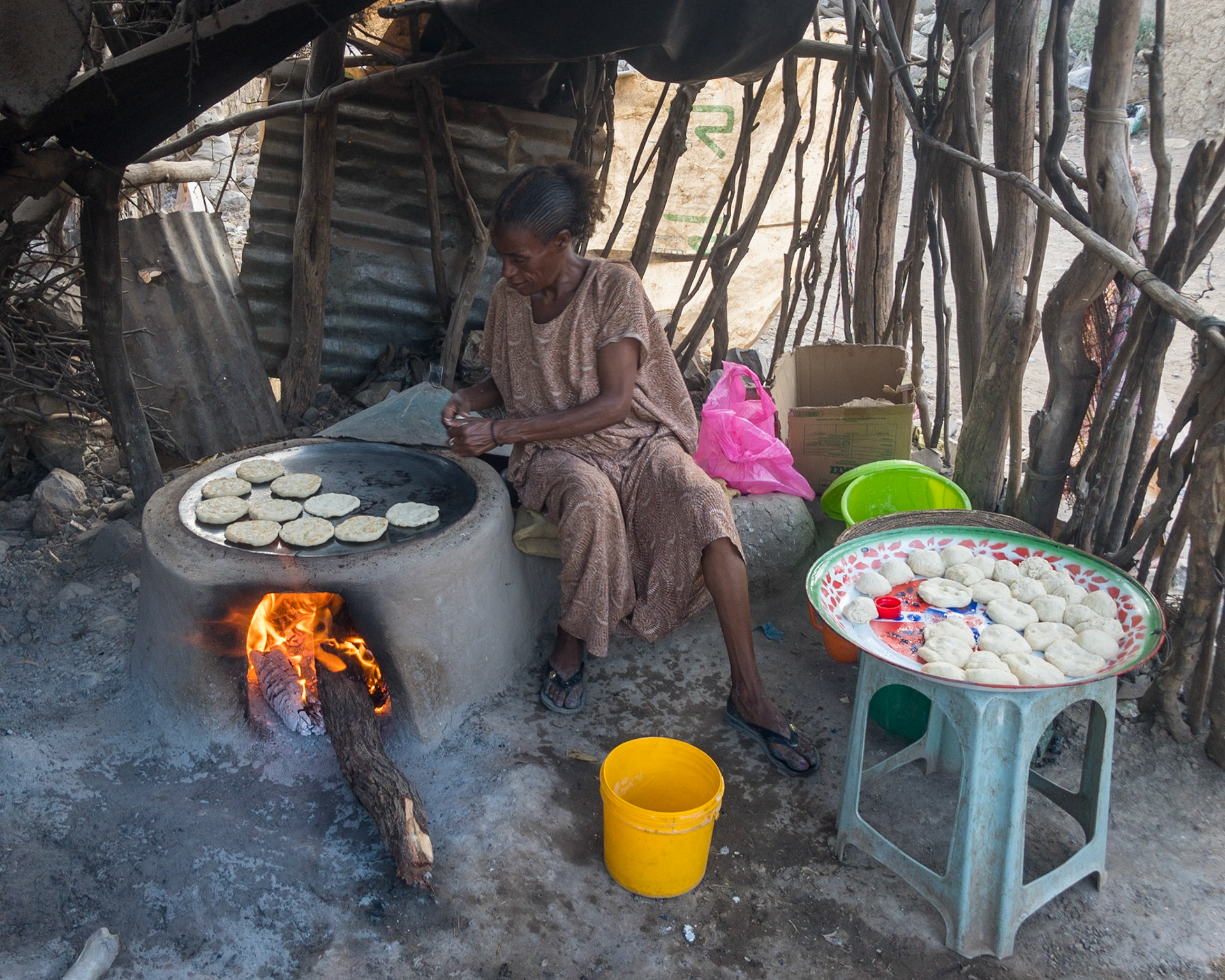 An Ethiopian woman makes and bakes small flat-breads over a flat iron-topped outdoor wood stove in a ramshackle enclosure of sticks, branches and corrugated iron sheeting.