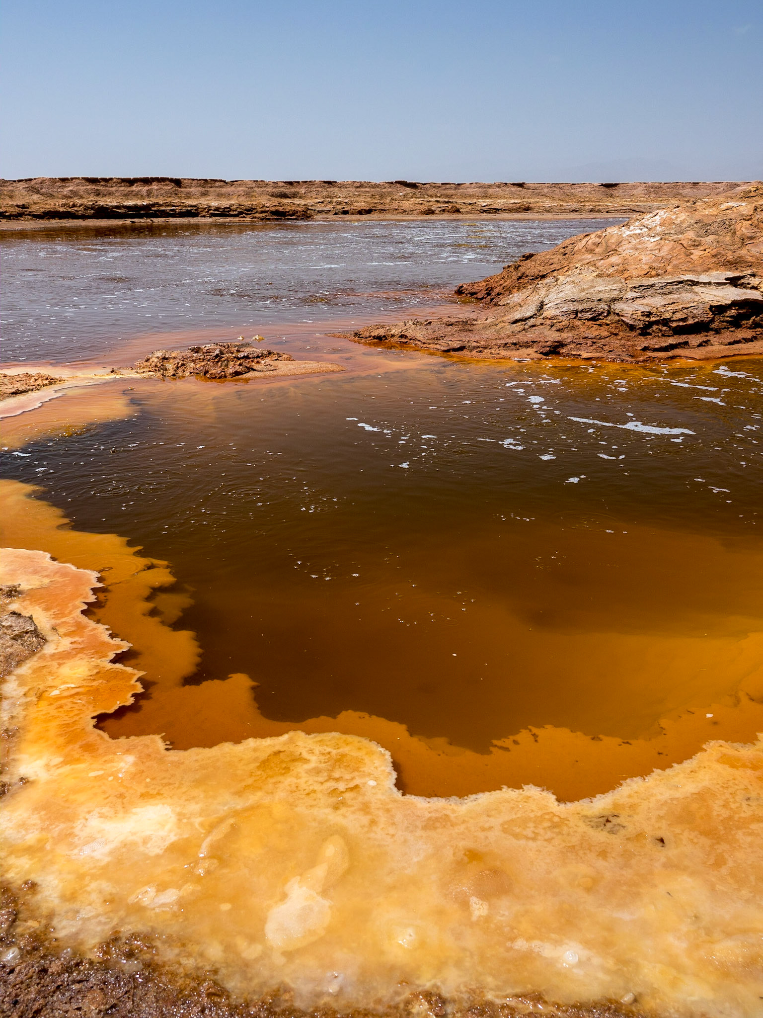 The clear, brown waters of a sulphuric acid lake bubble and churn creating orange-brown banks of salt on a flat landscape in the Danakil Depression, Ethiopia, the hottest, driest, most inhospitable place on Earth.