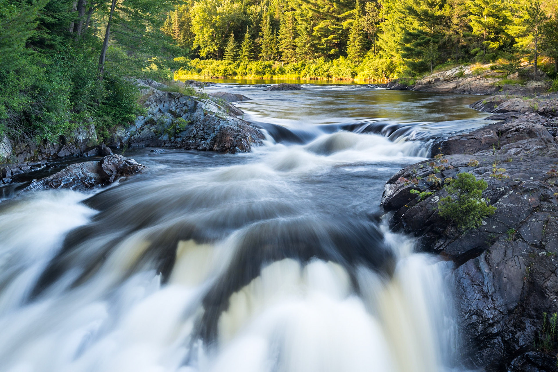 Waterfalls along a fast-flowing northern river tumble over the steep granite of the Canadian Shield along a forested riverbank lit golden by the evening sun.