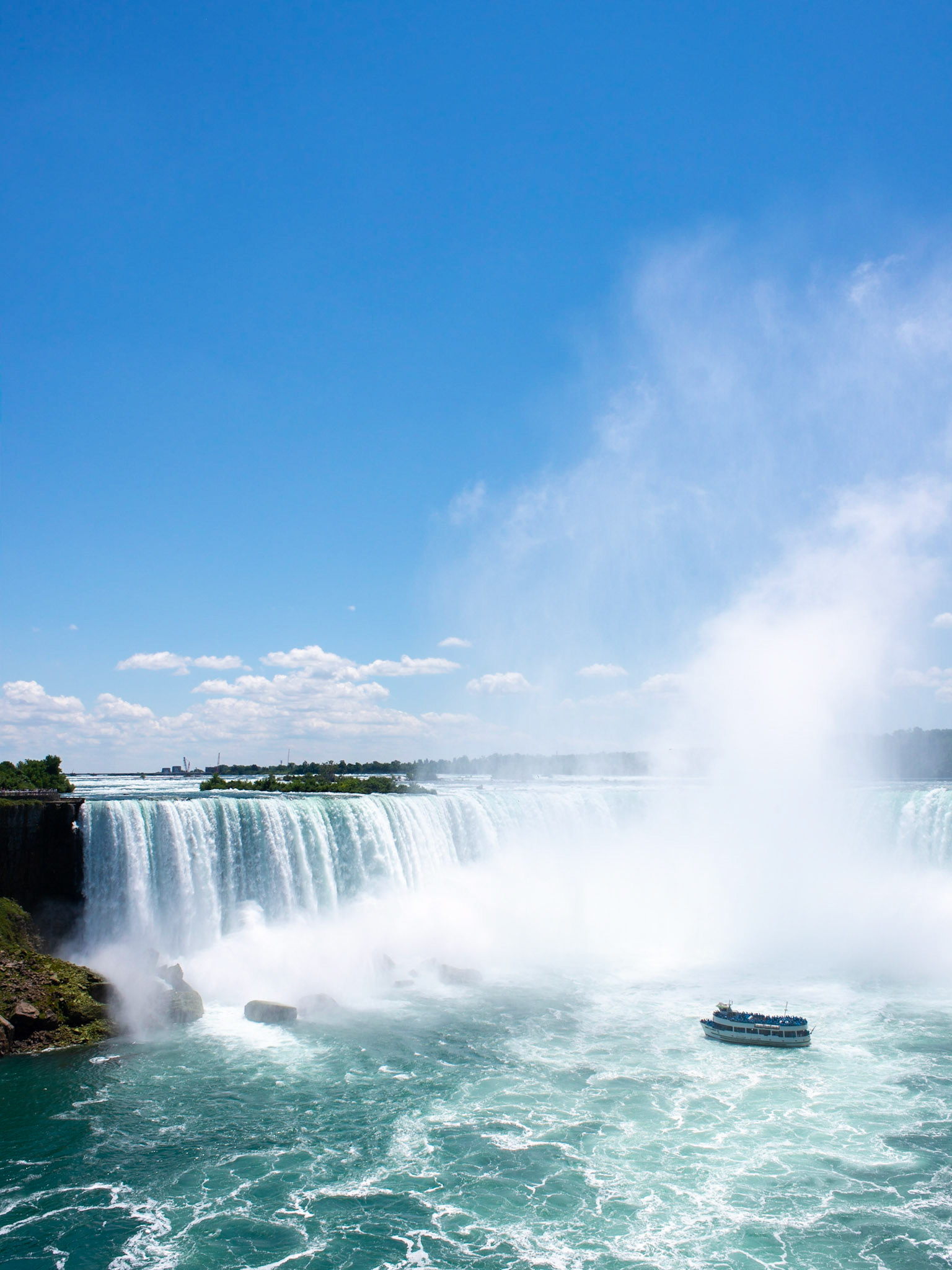 The Maid of the Mist tour boat approaches the base of the Canadian Horseshoe Falls of Niagara Falls amidst rising mist on a summer day with a bright blue sky along the Niagara Escarpment, a UNESCO World Biosphere Reserve in Ontario Canada