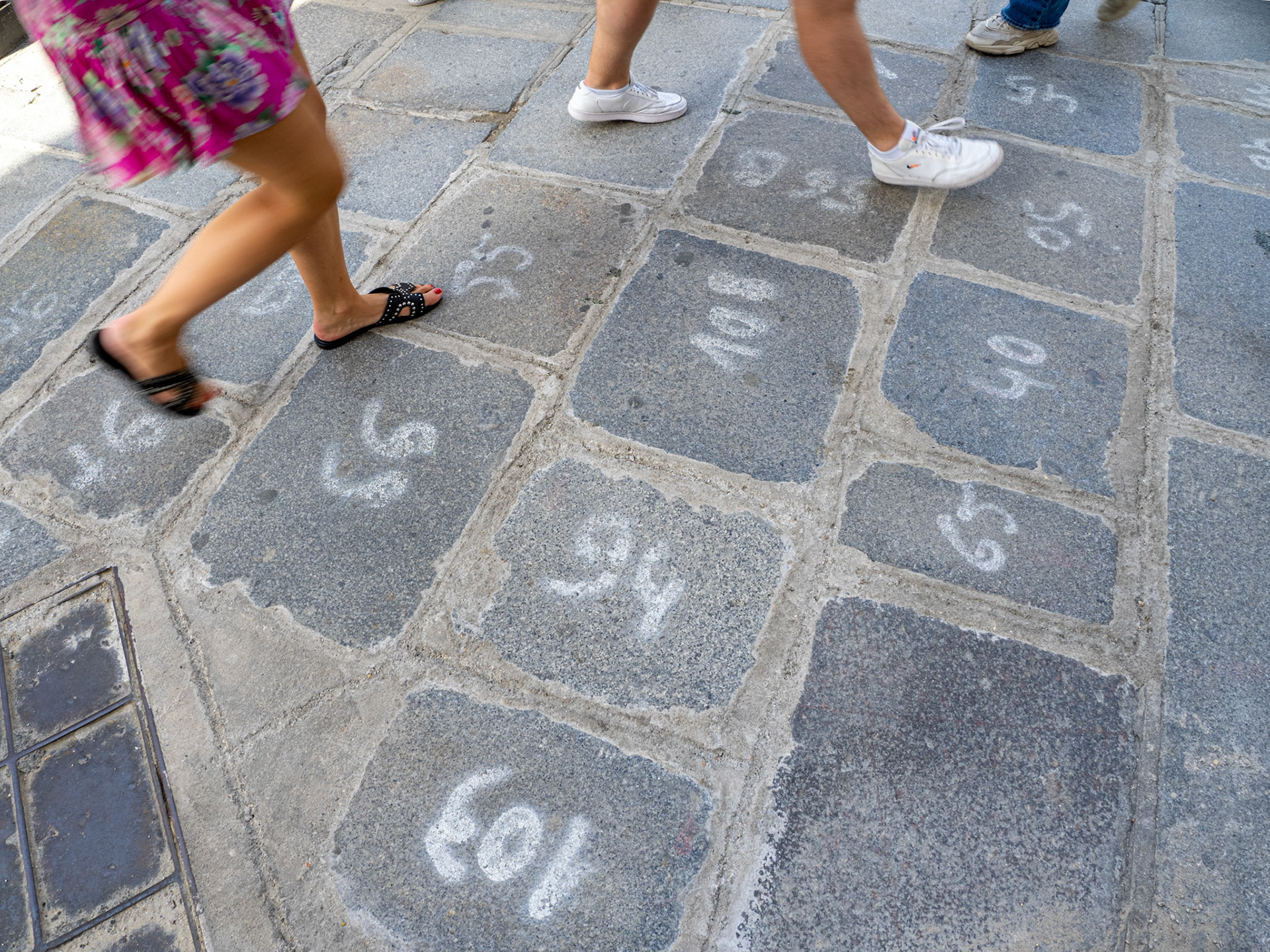 The feet and legs of passersby surround the numbered grey paving stones of a street in Paris.