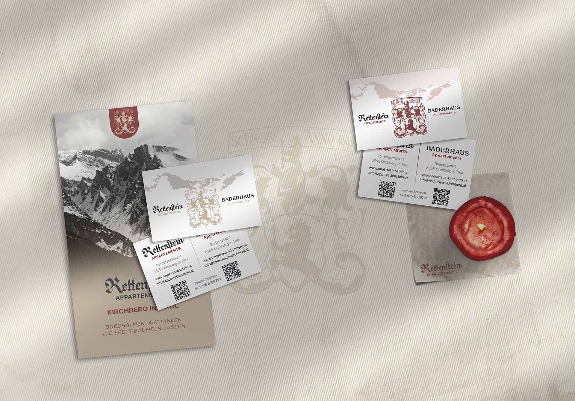 Visitenkarten und Flyer der Appartements Rettenstein und Baderhaus, in dunkelrot, gold und weiß, mit Logo und Kontaktinformationen. Beige Tischdecke, Serviette und Kerze im Hintergrund.