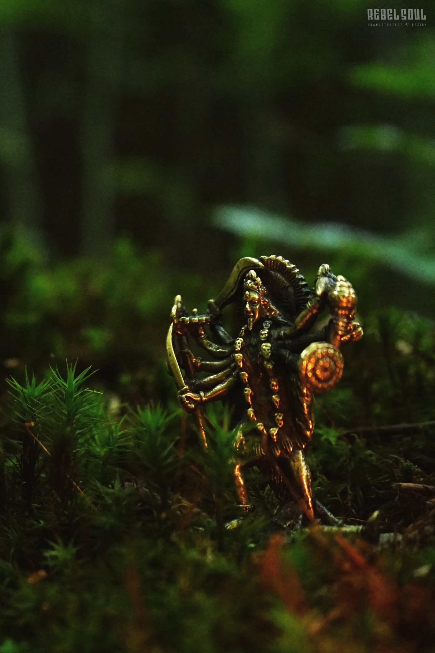 Makroaufnahme: Bronze-Kali-Statue im Moos, reflektierendes Licht von Feuer und Lichtung, im Hintergrund tiefes Grün des Waldes.