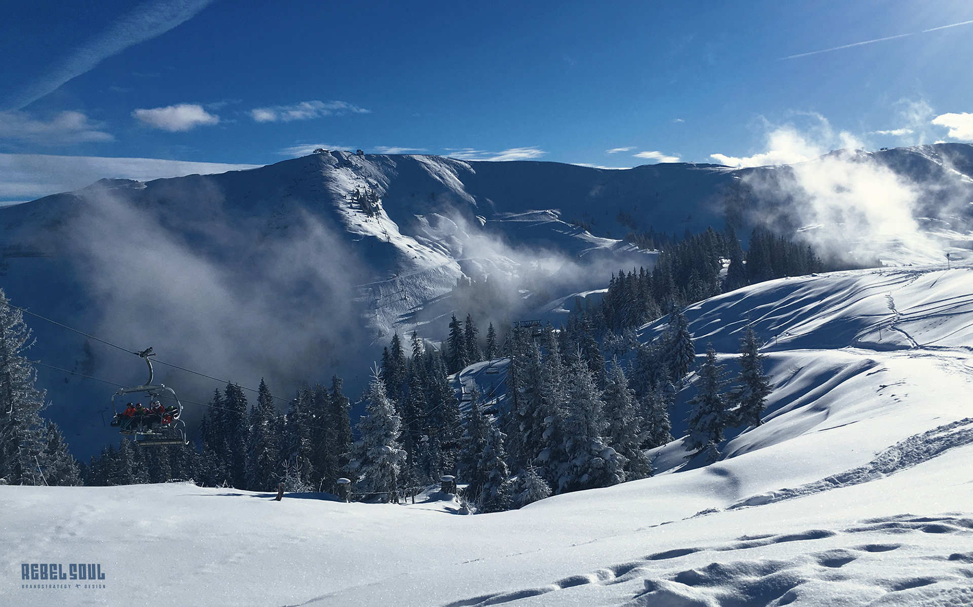 Kitzbüheler Alpen: Schneelandschaft mit schneebedeckten Nadelbäumen, Dunst, blauem Himmel und weißen Wolken. Gondel mit Skifahrern.