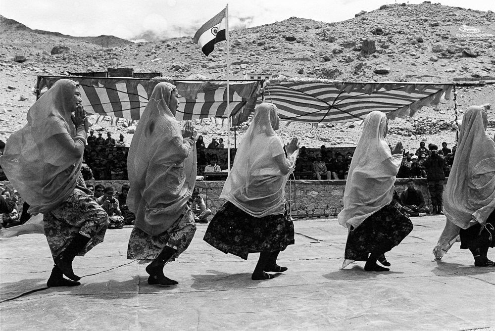A group of girls are performing national dances in front of a platform where local authorities are seated.
