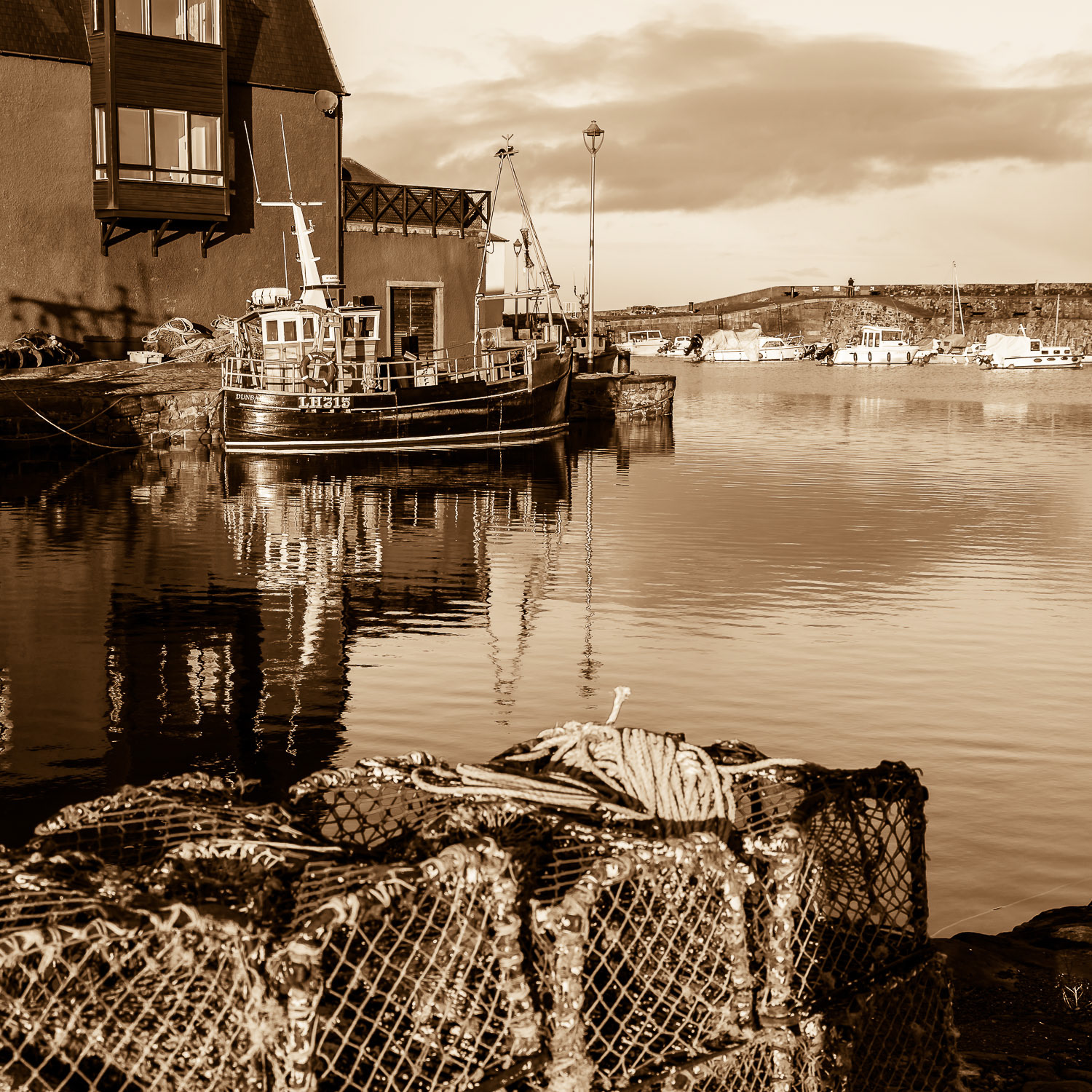 A monochrome split-toned image of a fishing boat tied up alongside the quay at the Cromwell Harbour, Dunbar, East Lothian