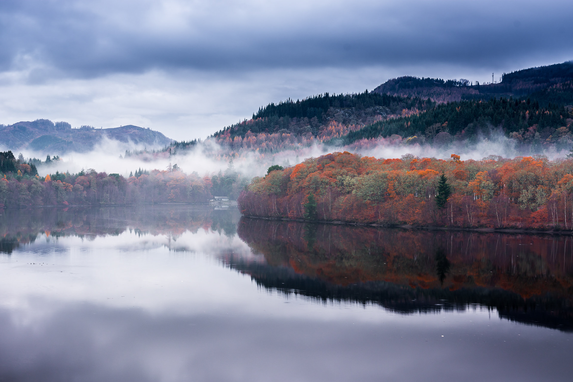 Mist hangs over the autumnal colours of the trees and the smooth surface of Loch Faskally, Perthshire, Scotland, with a remote cottage providing some scale and perspective to the scene