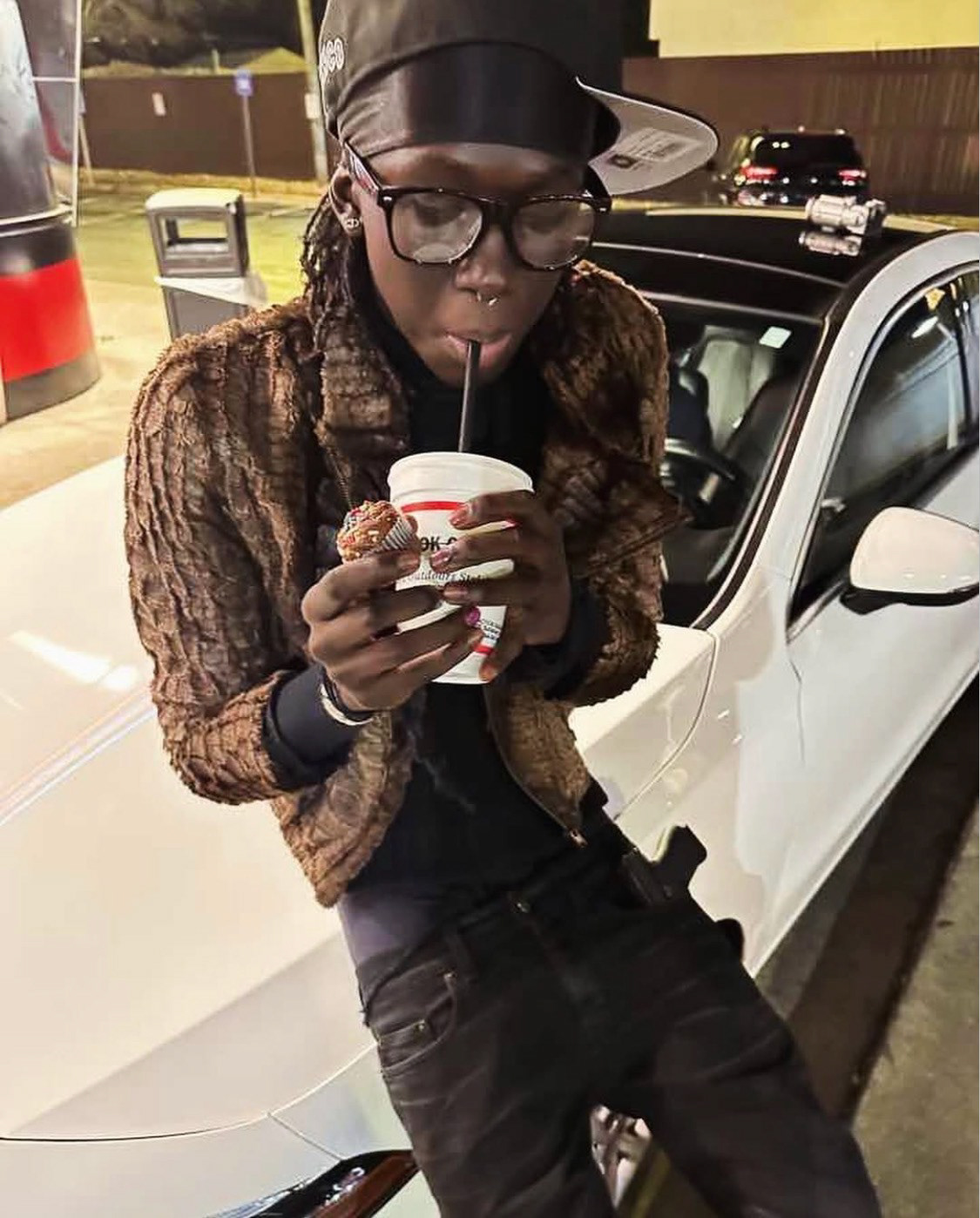 Payusu Njie (@p3ezy) at a gas station at night, leaning on a car. He wears a textured brown jacket, glasses, and a cap while drinking from a cup and holding a muffin.