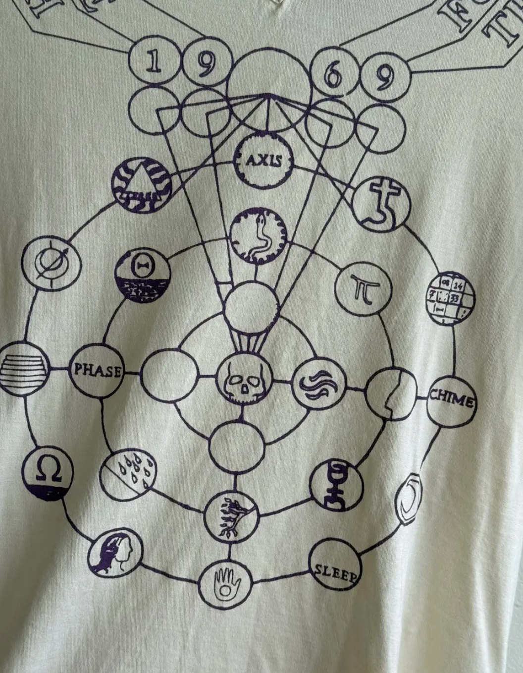 A close-up of a cream-colored t-shirt with a complex, dark purple graphic. The design is a circular, esoteric diagram featuring interconnected nodes, symbols (like a skull, hand, Pi, and Omega), and text elements such as "AXIS," "PHASE," and "SLEEP," with the numbers "1 9 6 9" at the top.