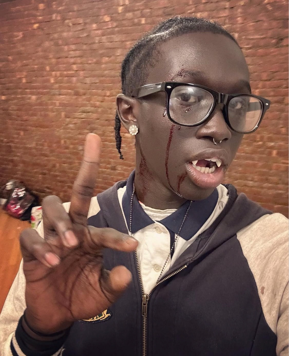 A close-up of Peezy in costume as a vampire, wearing plastic fangs and fake blood dripping from his eye and mouth. He wears large black-framed glasses, a nose ring, and a zip-up hoodie over a polo shirt.