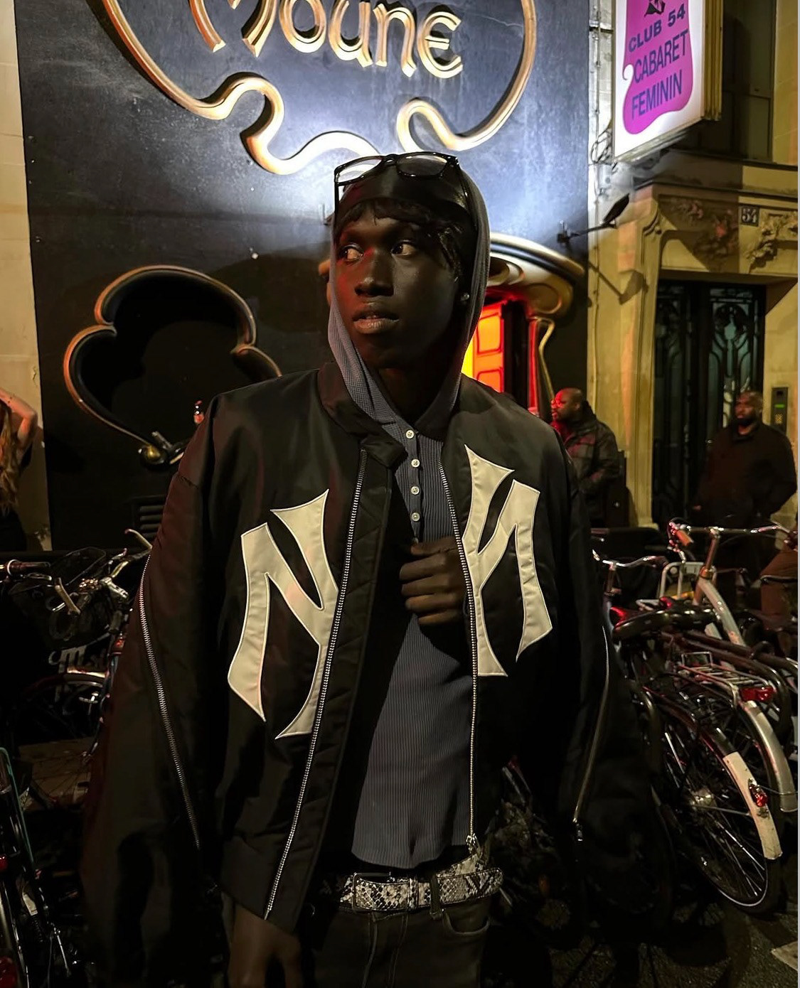 Payusu Njie (@p3ezy) at night in front of a club, wearing a black Mowalola "bootleg" New York Yankees bomber jacket over a grey hoodie and pinstriped shirt.