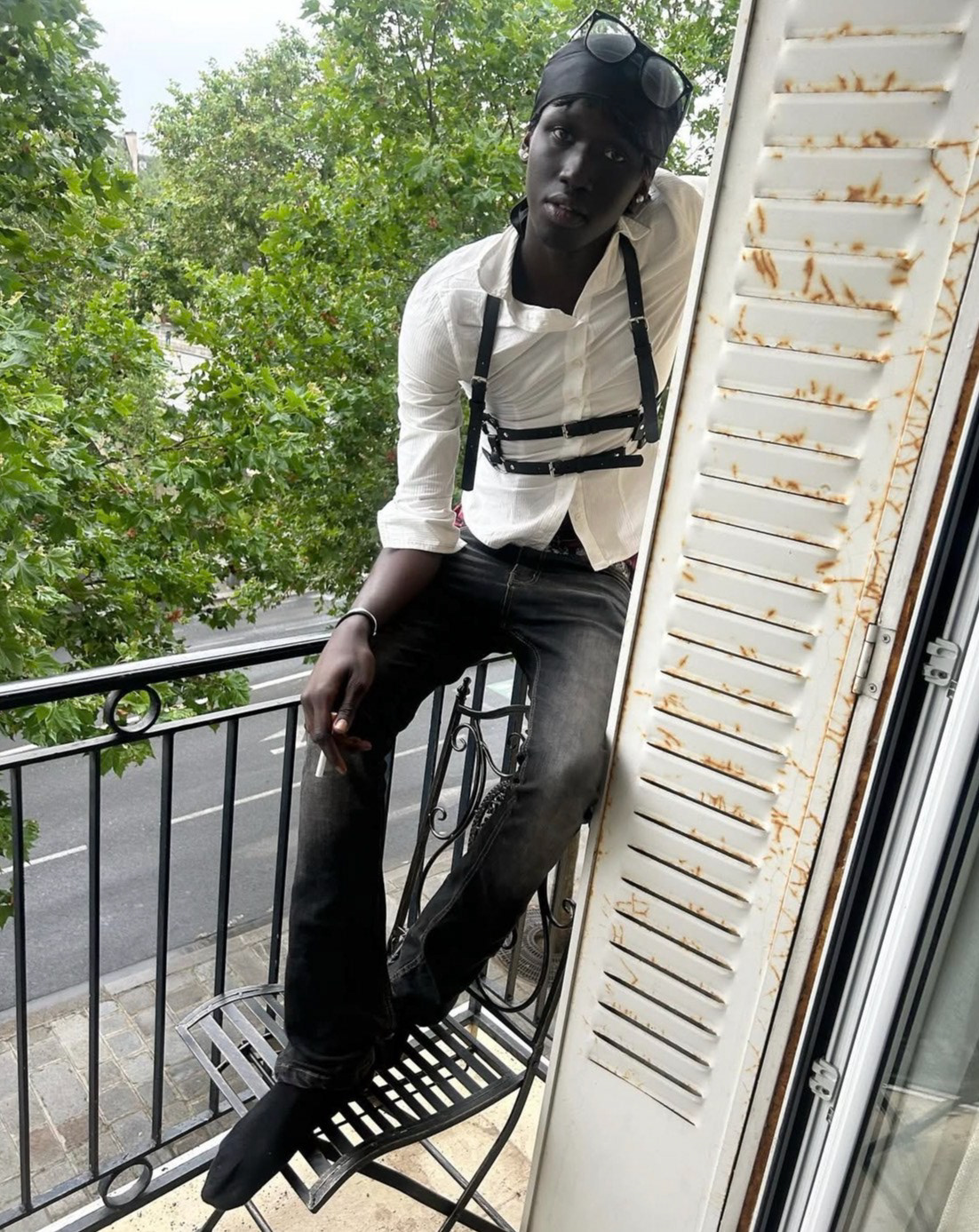 Payusu Njie (@p3ezy) posing on a balcony, wearing a white button-down shirt accessorized with a black leather body harness. He wears a black durag, glasses, and faded black jeans.