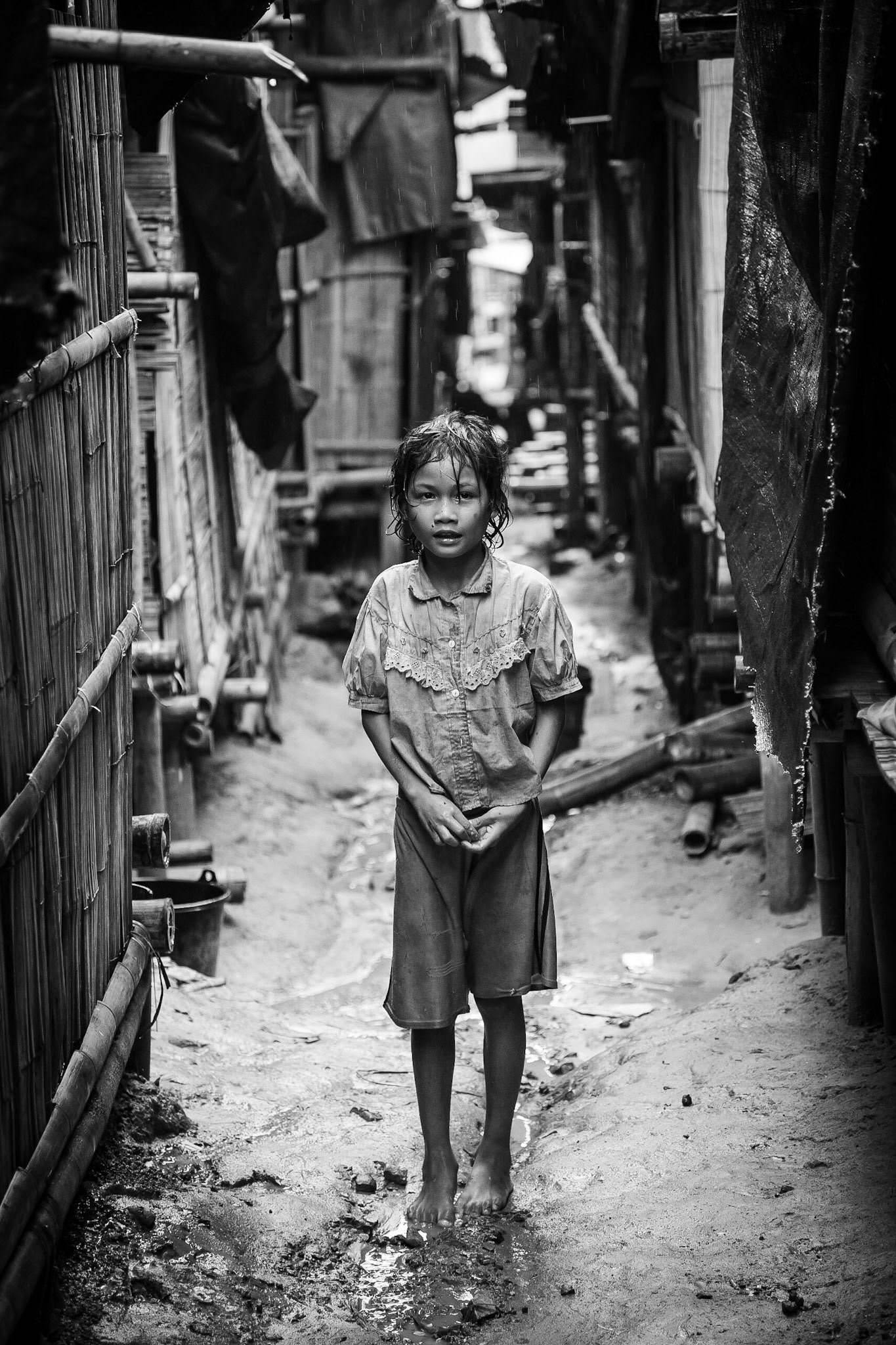 A Burmese Karen refugee child standing barefoot in the rain in a narrow street between the bamboo shelters.
