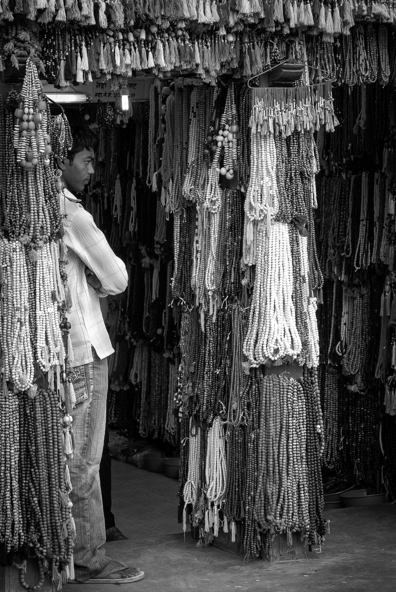 A vendor of Budhist prayer beads looks out from the door way of his shop surrounded by strings of beads.