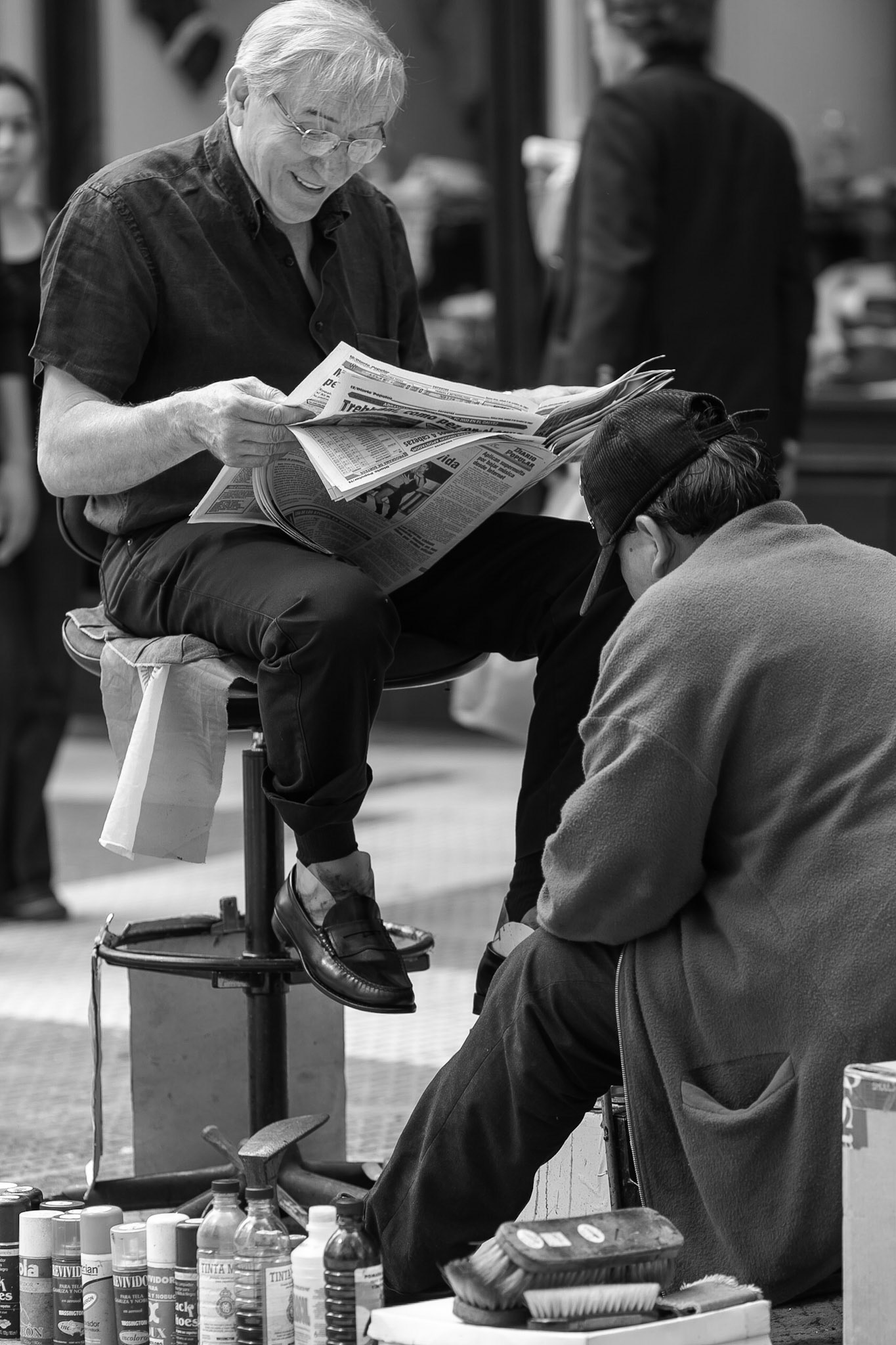 Man receives a show shine on the street in Buenos Aires, while seated and reading a newspaper.