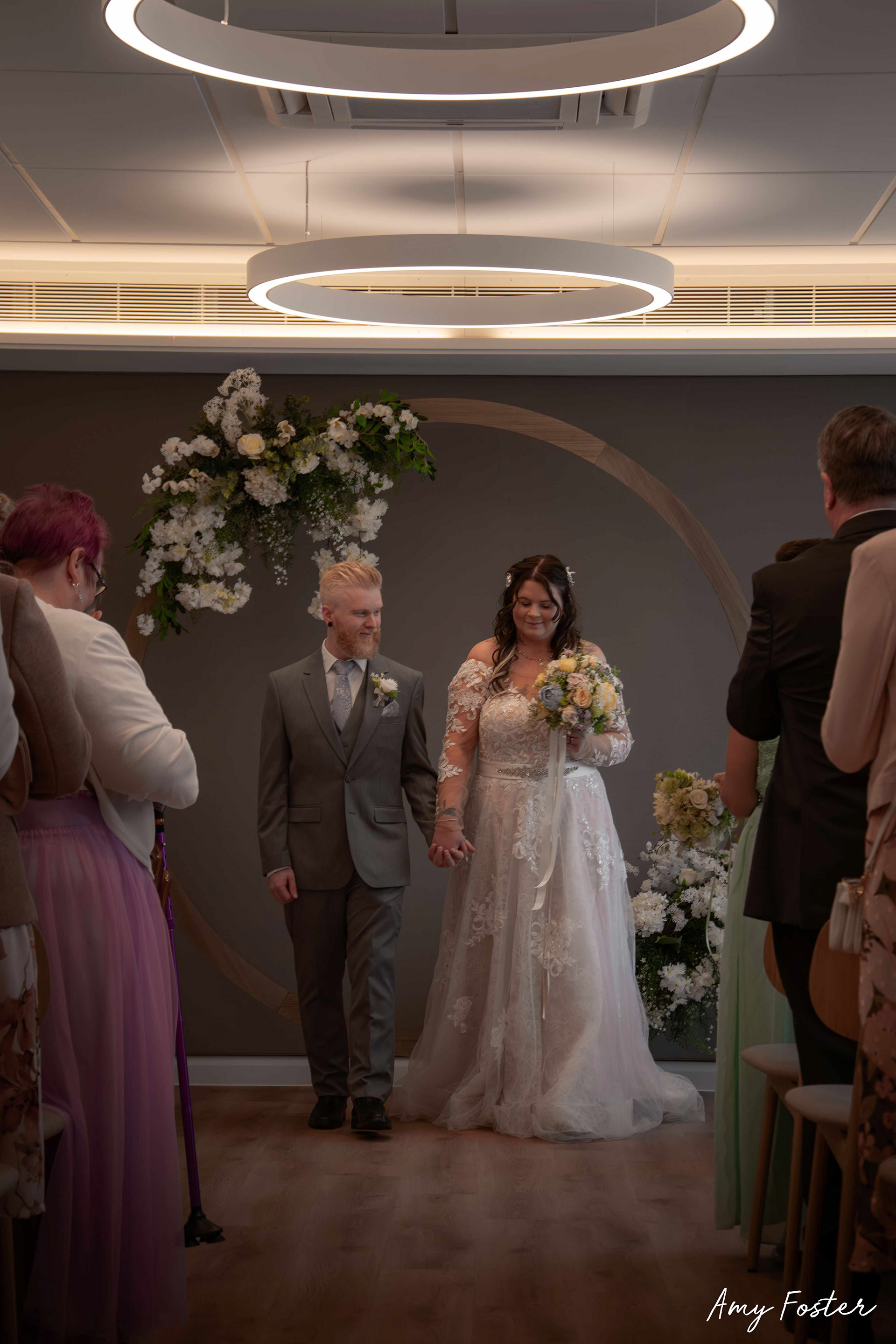 Bride and groom holding hands leaving the ceremony together, guests line the edges of the frame and the couple are smiling