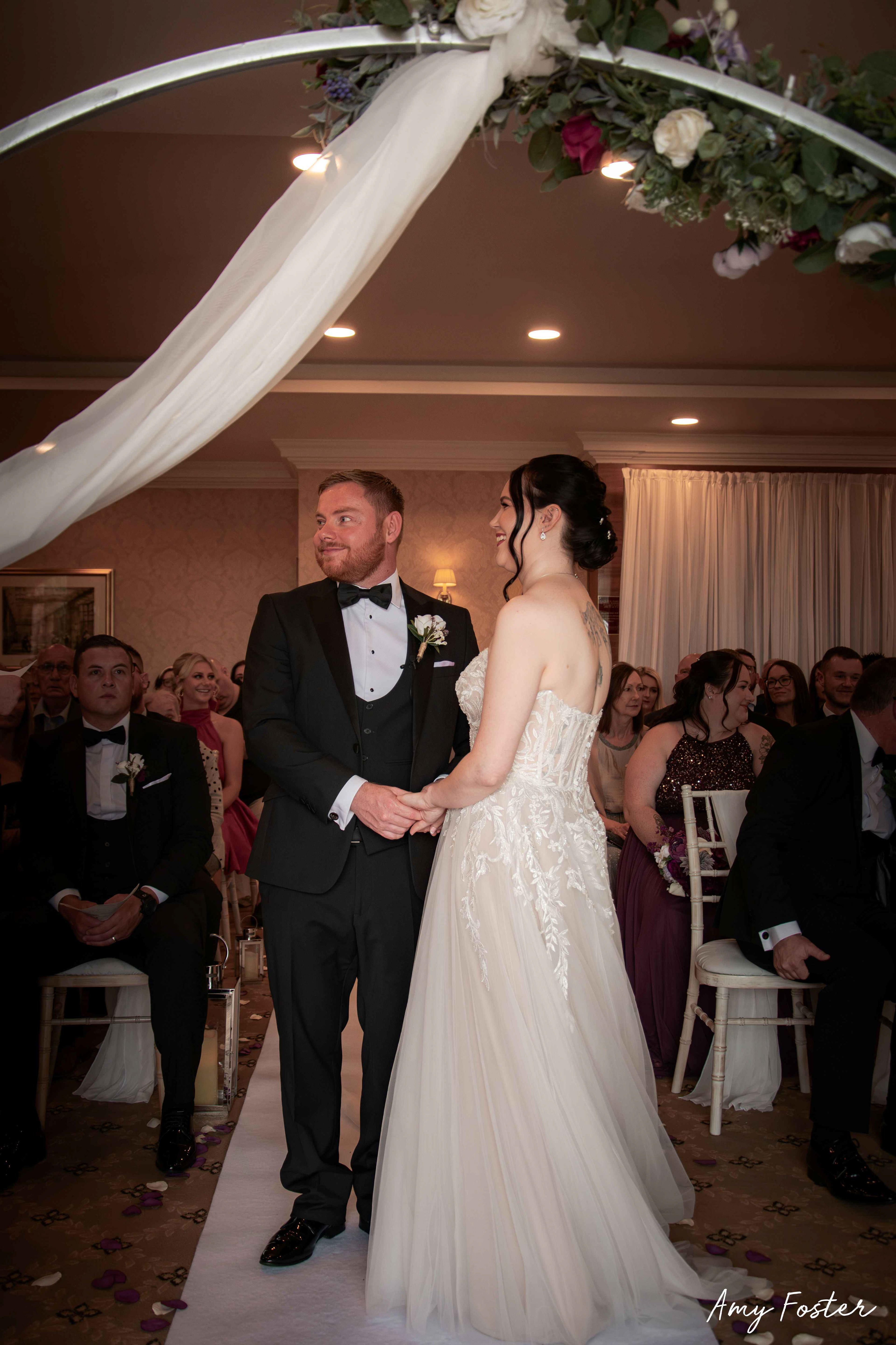 Couple holding hands smiling, full body photos under archway of flowers at the end of the isle