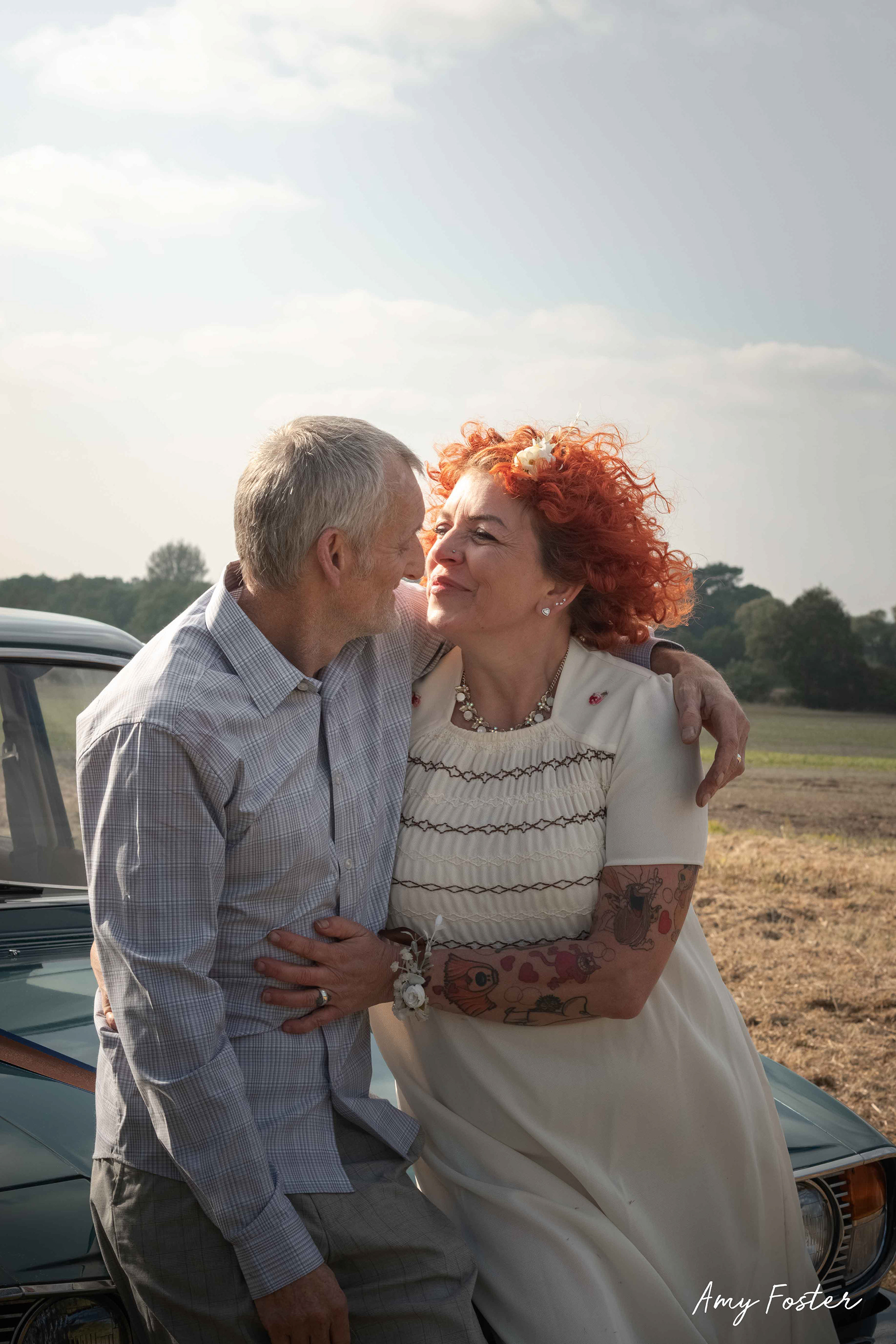 Newlyweds sat on retro car bonnet faces close smiling at one another on a summers day 