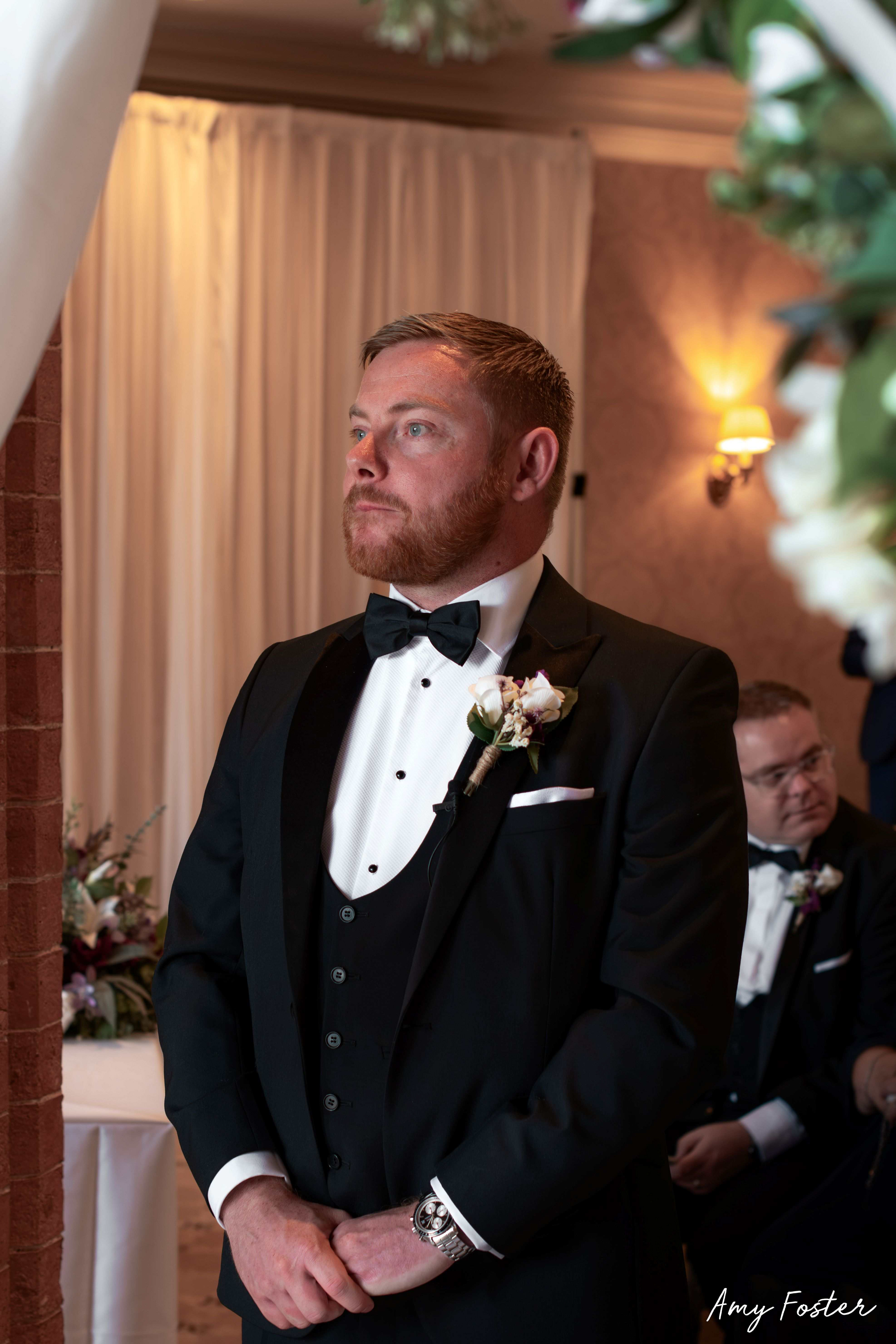 groom in black suit waiting for bride at the end of the isle framed in flower archway  