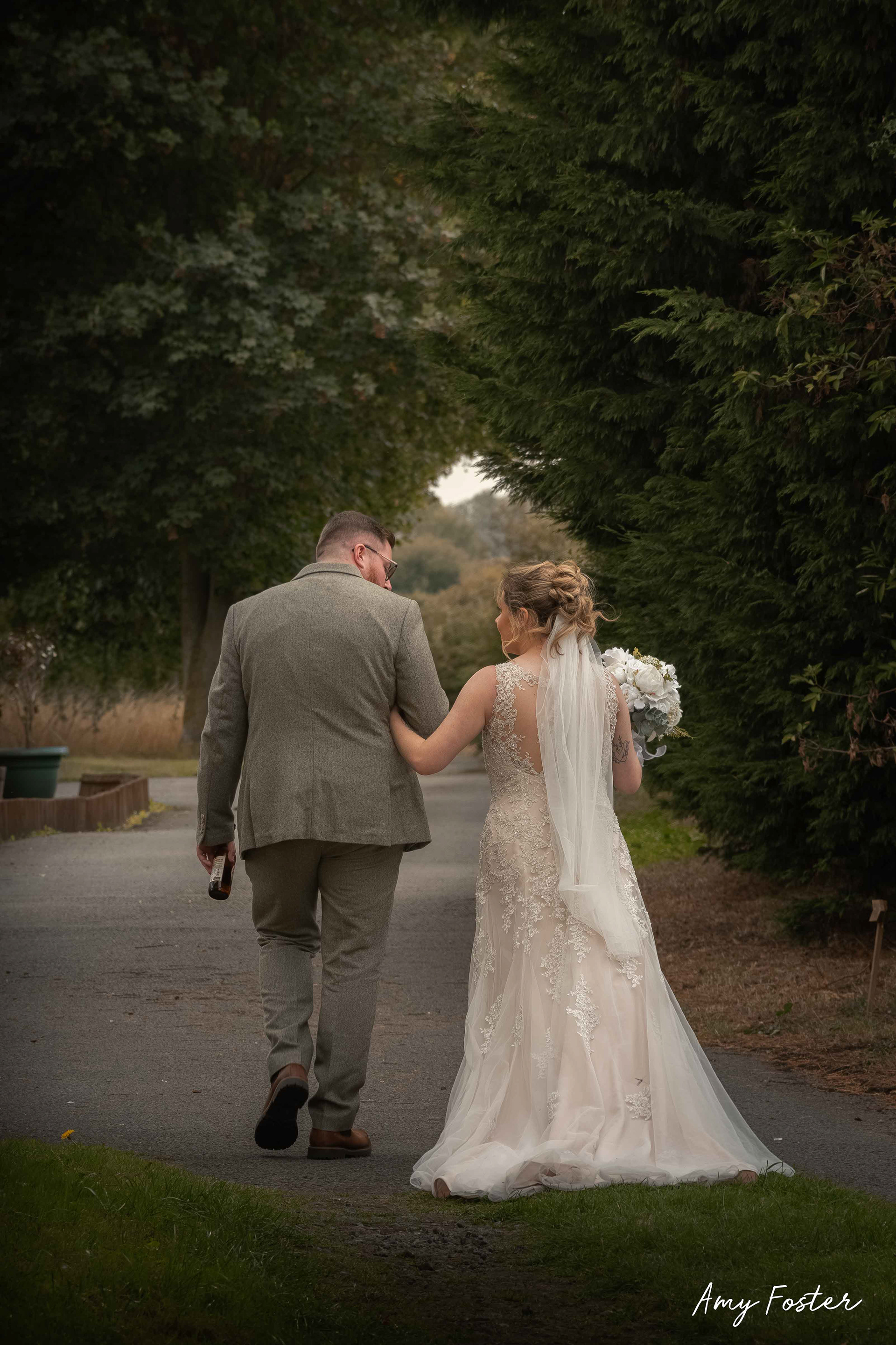 Bride and groom walking arm in arm away together looking at each other down an outdoor tree lined path