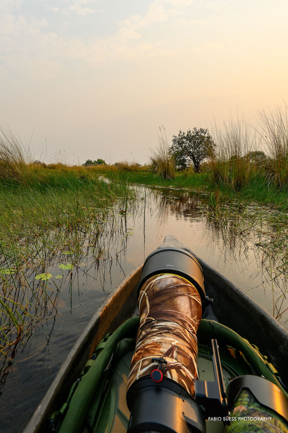 Fabio Süss im traditionellen Mokoro bei Sonnenaufgang im Okavango Delta, mit Teleobjektiv bereit für Wildlife-Fotografie, umgeben von stiller Natur.
