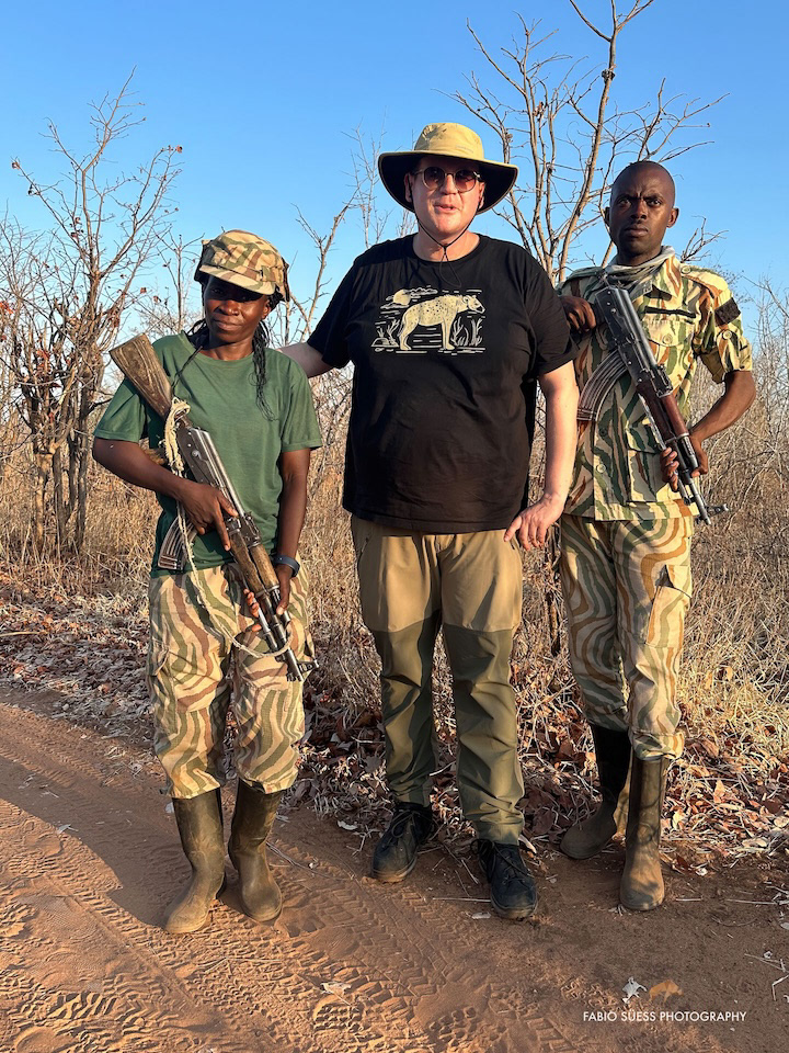 Fabio Süess steht in der Mitte zwischen zwei bewaffneten Wildhütern in Tarnkleidung in einer trockenen Buschlandschaft in Afrika. Er trägt einen Sonnenhut, Sonnenbrille, ein schwarzes T-Shirt mit Tiermotiv sowie beigefarbene Outdoor-Hose. Die Szene wirkt ruhig und zeigt Respekt gegenüber der Arbeit der Ranger.