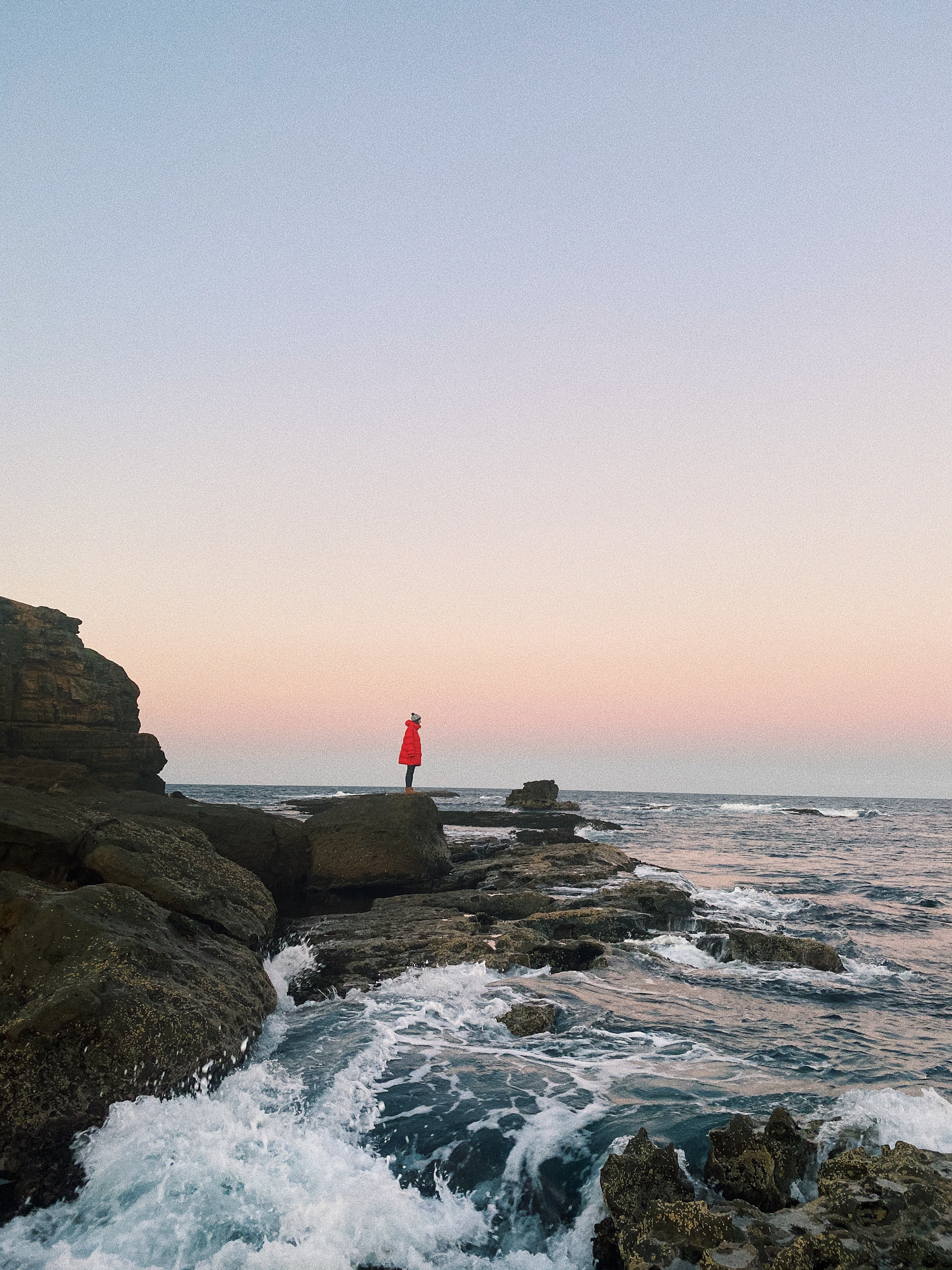 woman with red puffer jacket, standing on rocks, overlooking the ocean.
