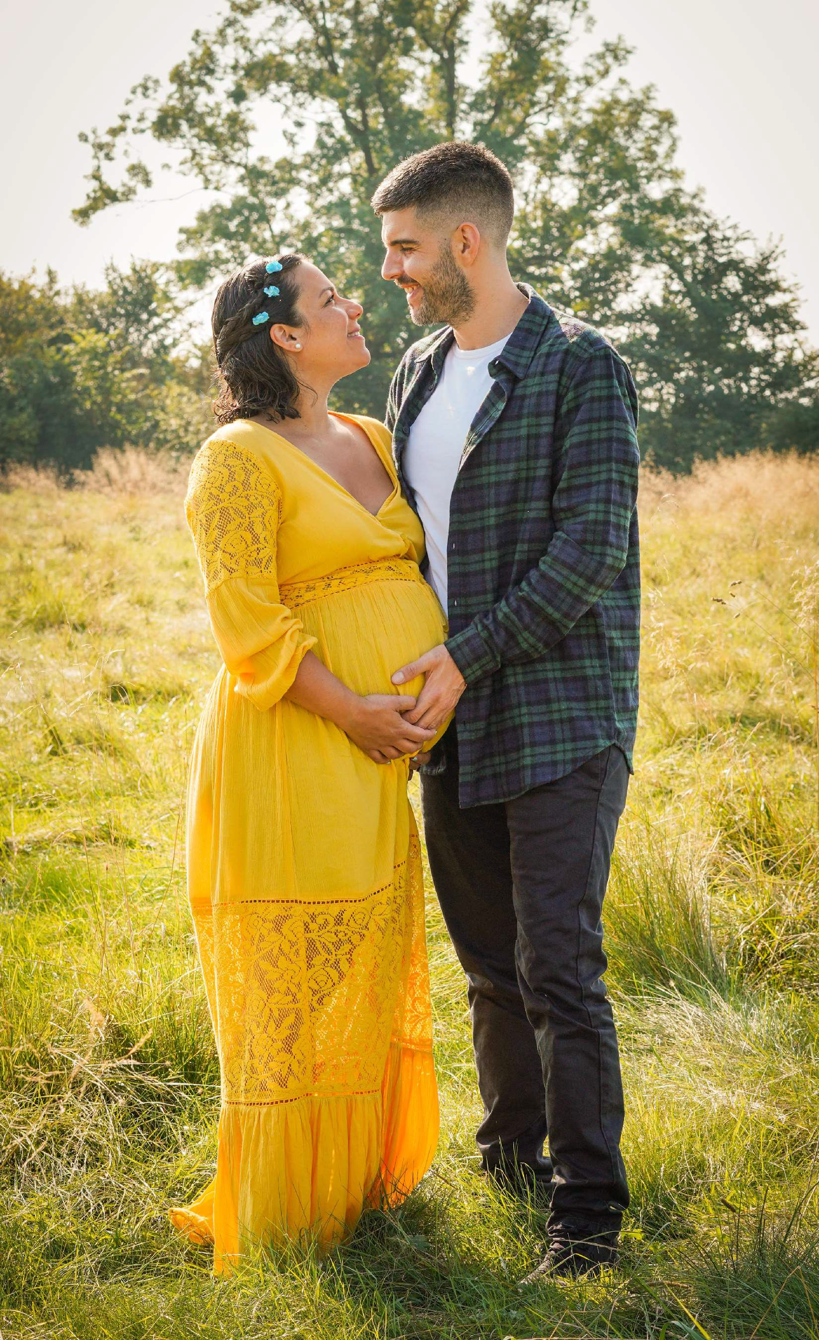 italian and greek couple stood holding each other on sunny day near steeple claydon in buckinghamshire smiling and holding bump on maternity photo shoot with david lefebvre photographer near buckinghamshire