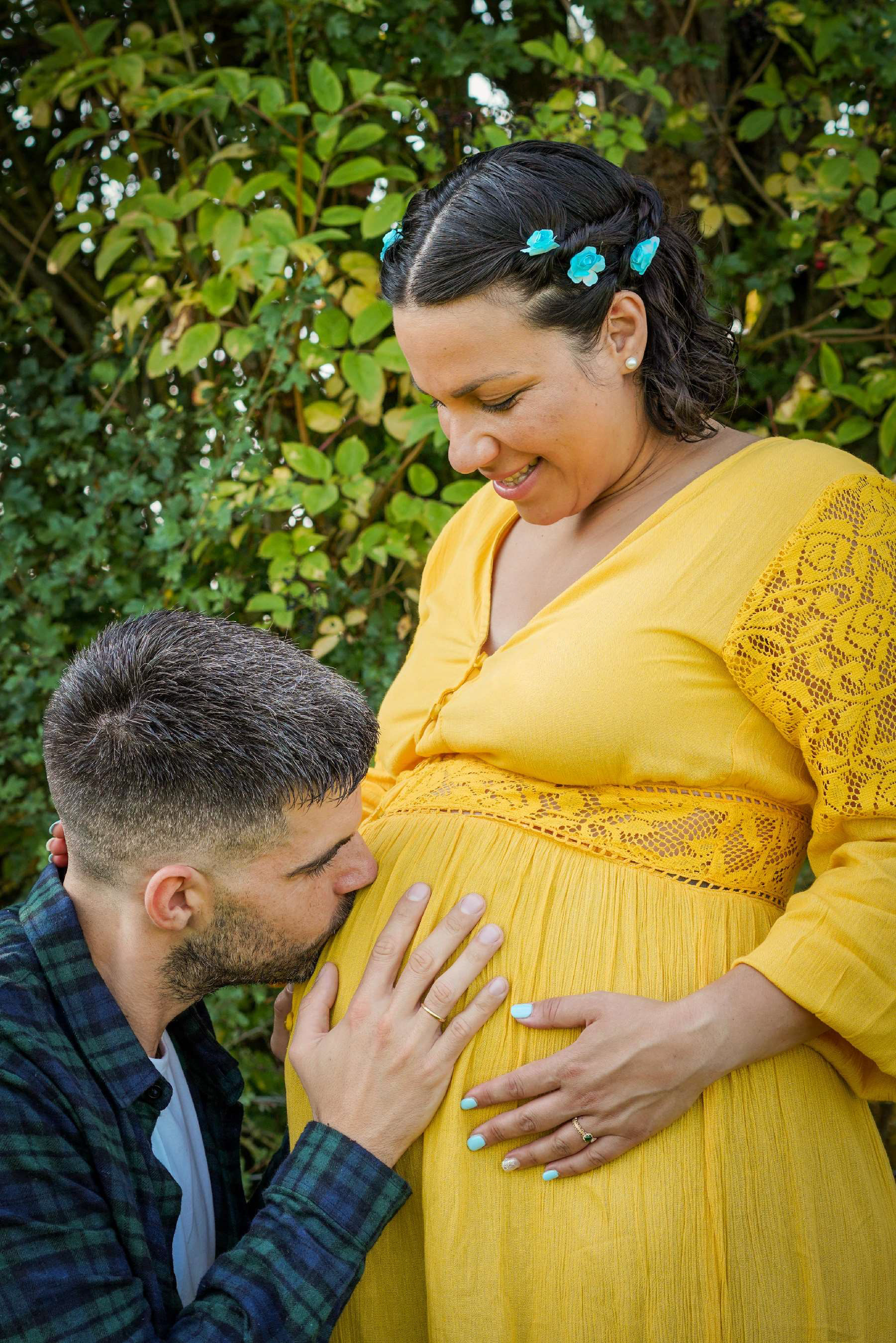 cute romantic photo of italian and greek couple stood together whilst he kisses her bump with a green background she is wearing a yellow maternity dress on photo shoot session with david lefebvre near aylesbury taken at claydon house