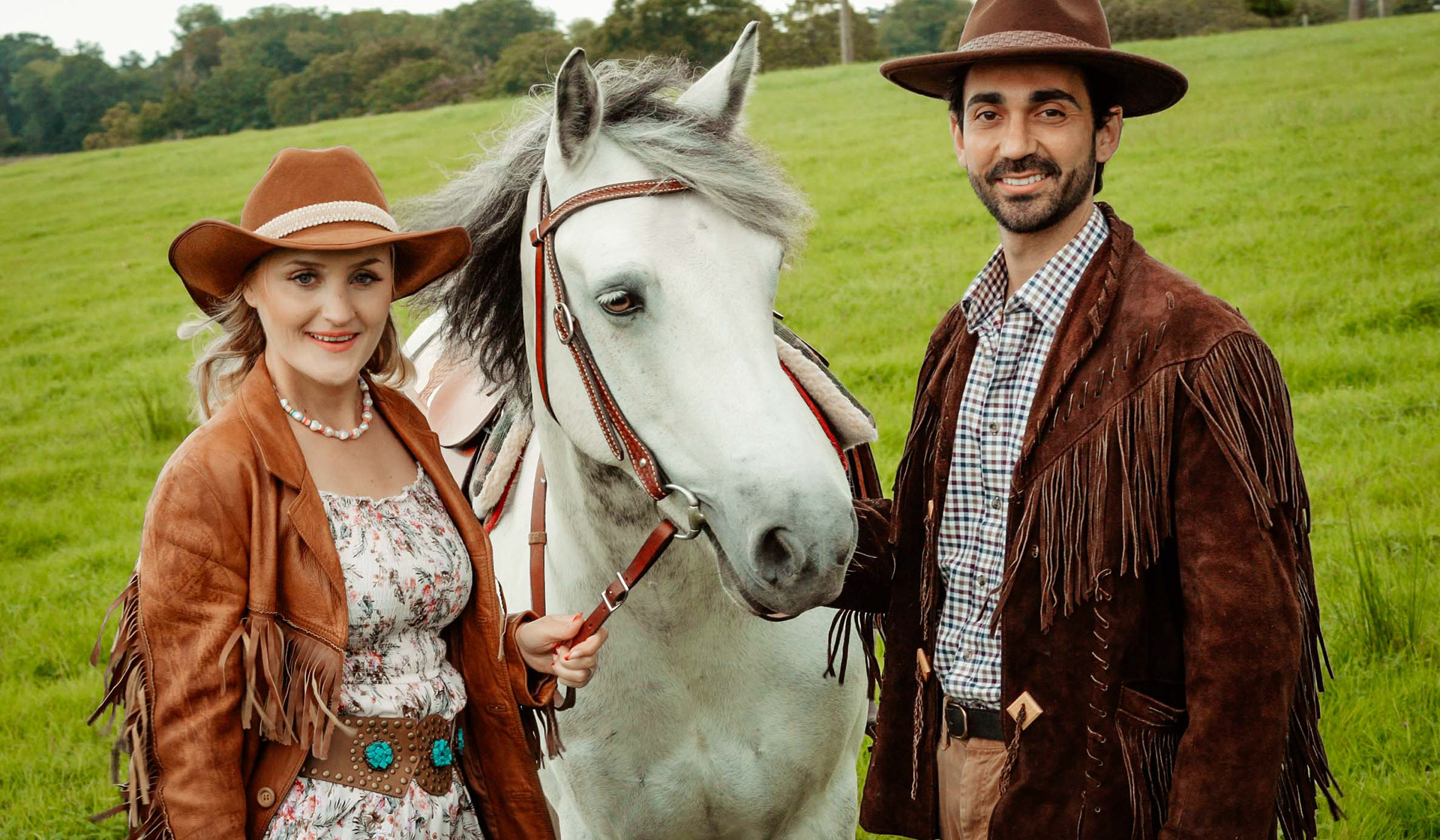 bride and groom smiling together with white horse wearing boho style cowboy jackets and hats in green field in sussex taken by wedding photographer david lefebvre from bicester and buckinghamshire