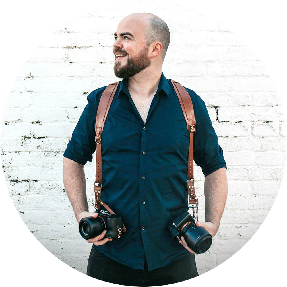 wedding portrait and product photographer david lefebvre from buckinghamshire stood in blue shirt with sony cameras and brown leather harness on white brick wall