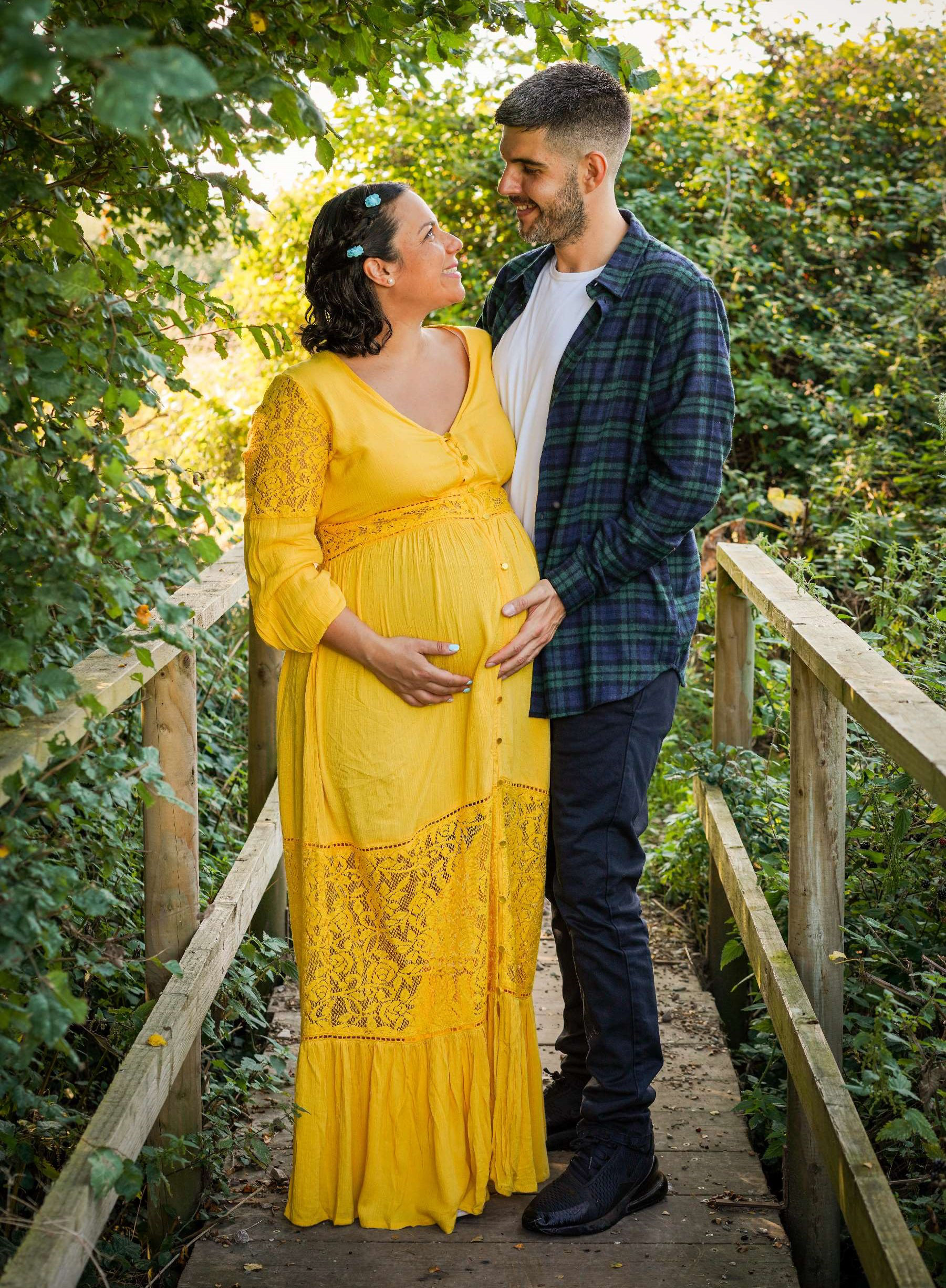 good looking greek couple leave kind words and review stood on wood bridge over stream in sunlight smiling wearing checkered shirt and long yellow dress on maternity shoot with steeple claydon photographer david lefebvre