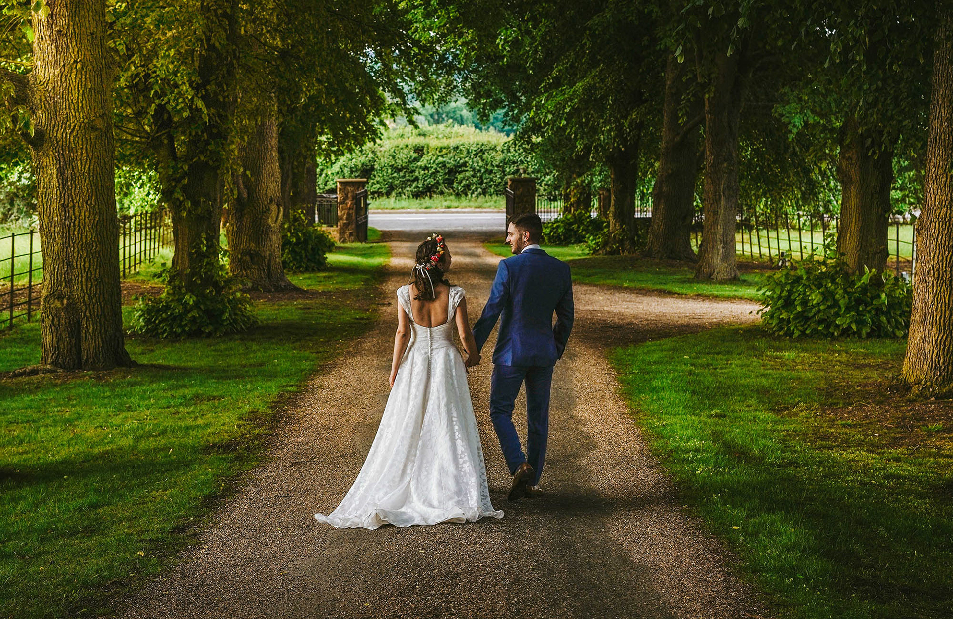 stunning golden hour image of bride and groom walking away from wedding photographer david lefebvre photography down dirt track driveway between huge trees at plum park manor in towcester northamptonshire
