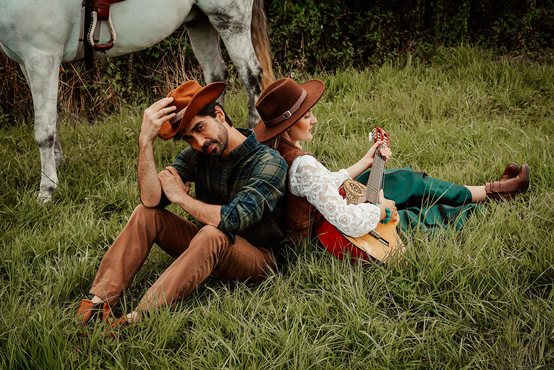 candid model sat with mexican cowboy husband smiling with white horse leaning on partner shes wearing and emerald dress and leathers holding accoustic guitar taken by aylesbury photographer david lefebvre