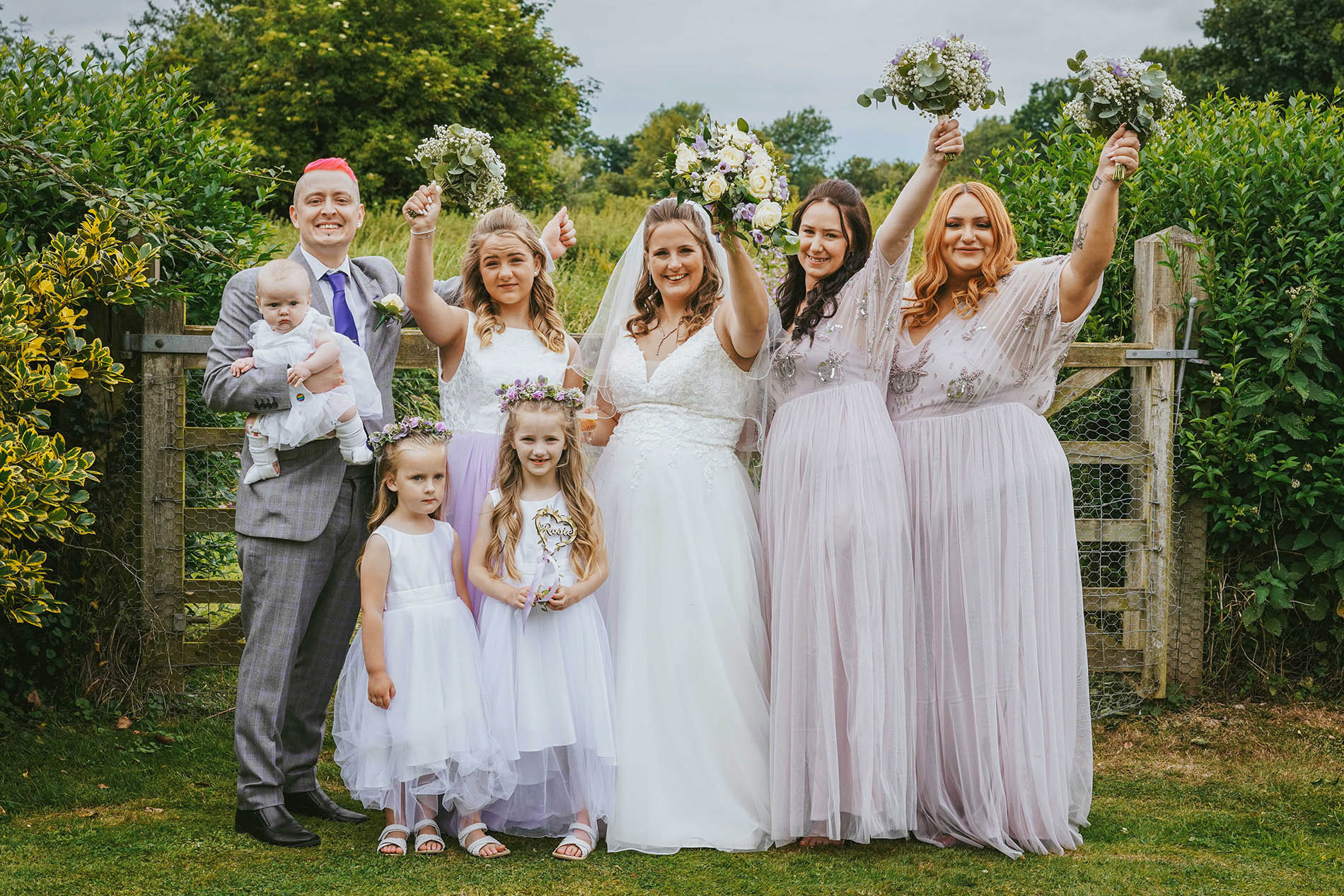 bridal party smile and have their colourful rose bouquets up in the air with pink red and blonde hair in lilac dresses taken outside manor farm barn by wedding photographer david lefebvre photography