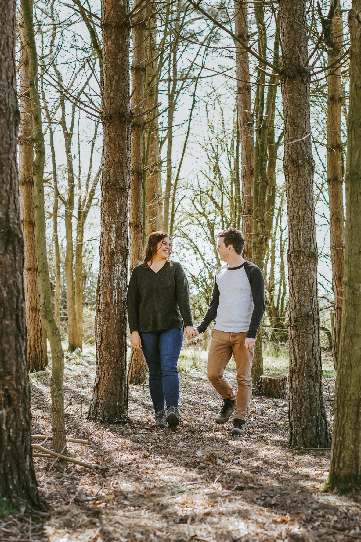smiling couple walking through sunny forest looking at each other whilst photographer david lefebvre takes their photos during candid pre wedding engagement shoot session
