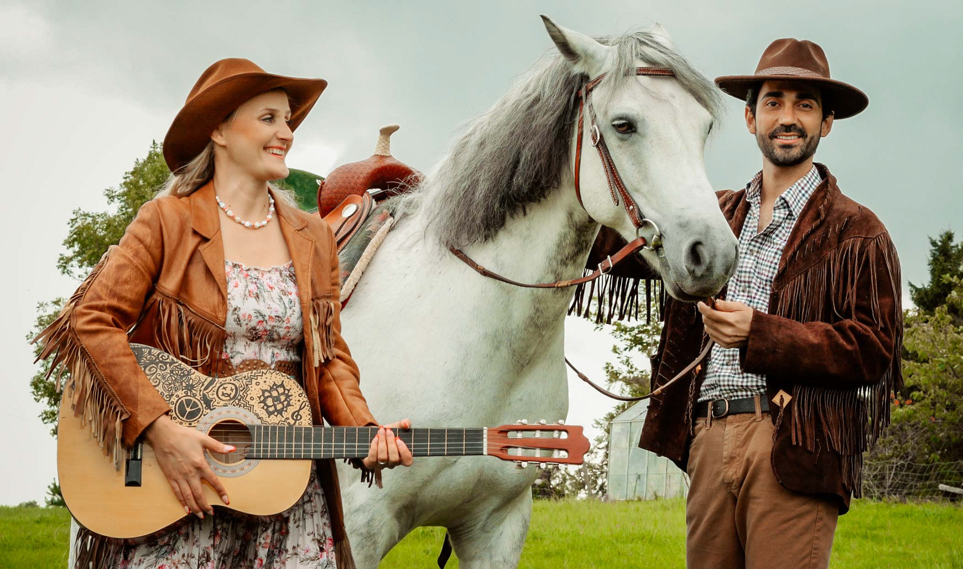 bride and groom smiling together with white horse wearing boho style cowboy jackets and hats in green field in sussex taken by wedding photographer david lefebvre from buckinghamshire