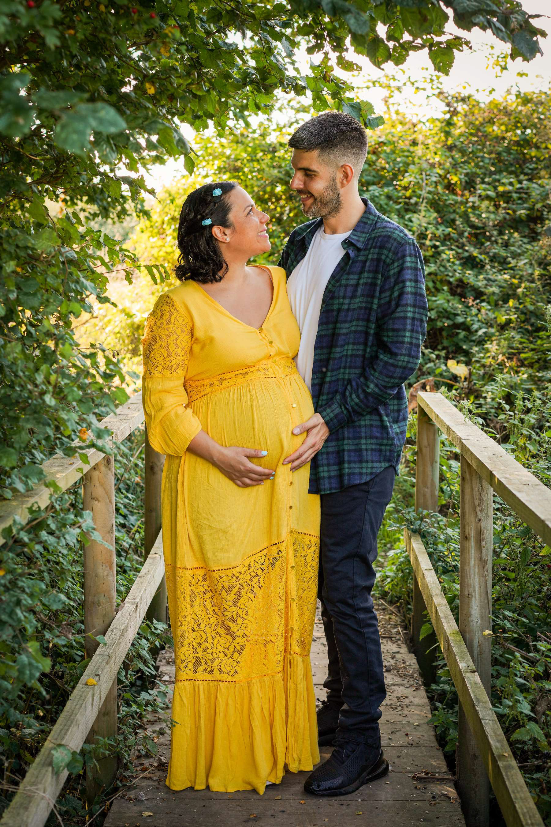 loving romantic italian and greek couple stood on bridge in warm sunshine surrounded by blackberry and wild berry bushes in with warm sunshine at claydon house near steeple claydon in buckinghamshire taken on maternity shoot by photographer david lefebvre
