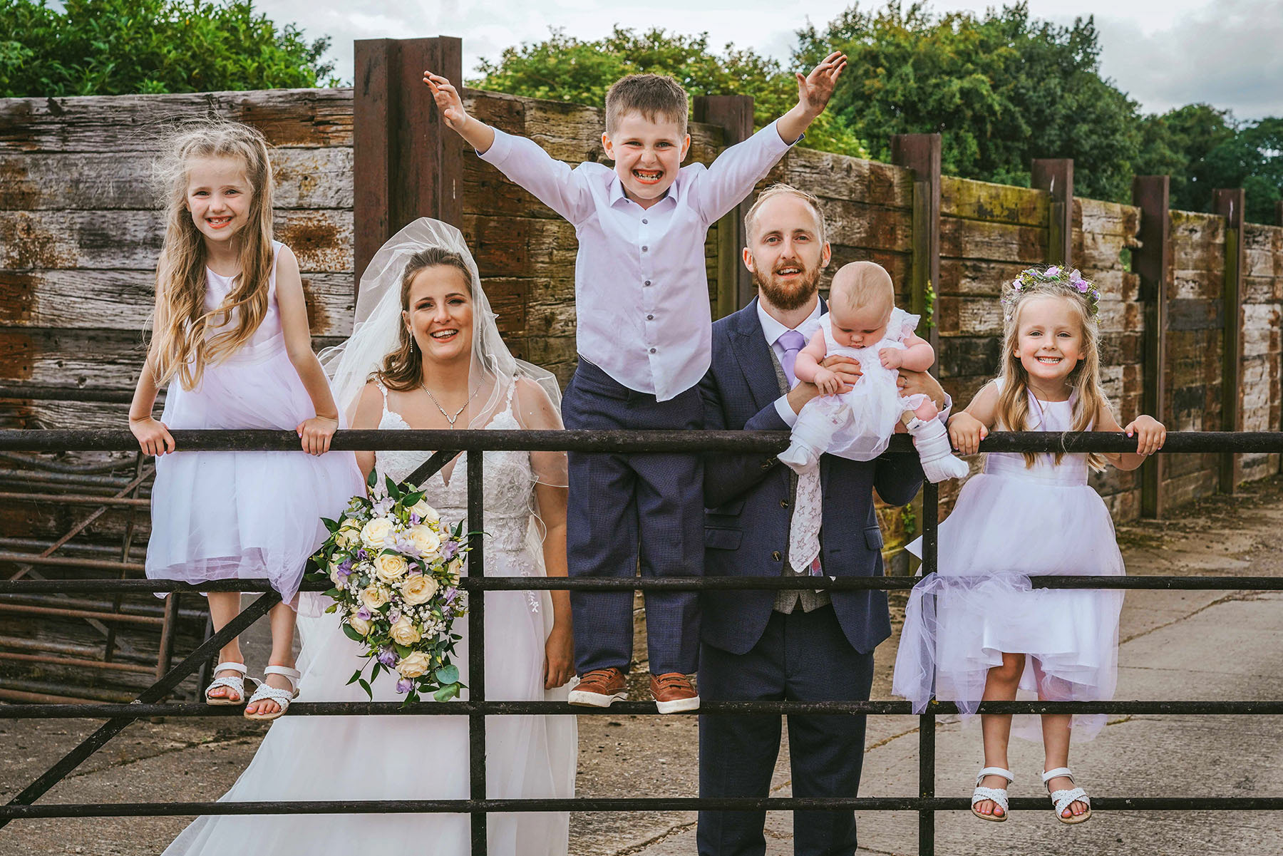 young smiling children stood on cattle gate with bride and groom outside at rustic manor farm barn wedding venue in marsh gibbon taken by david lefebvre wedding photography