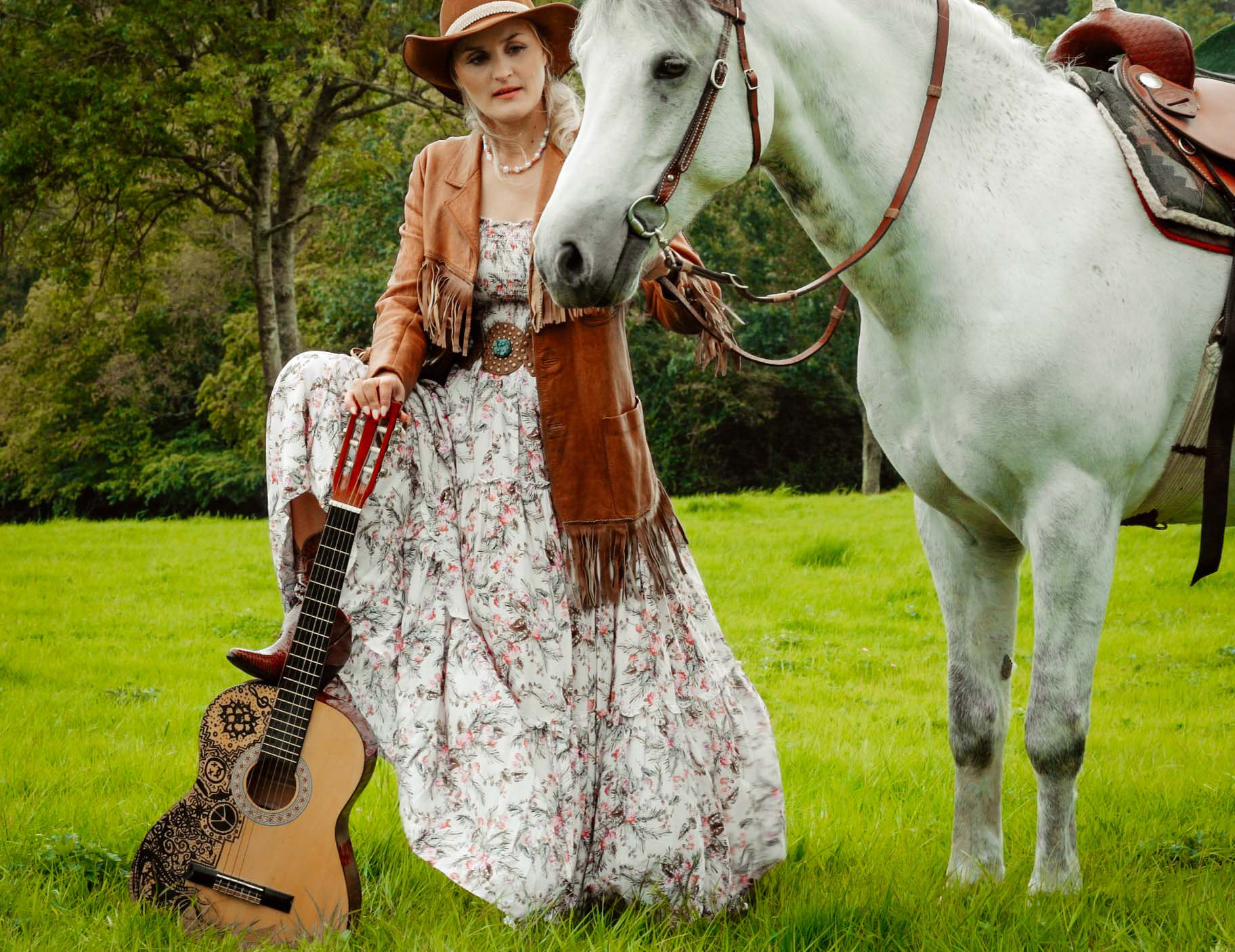 bride stood in floral dress leaning on guitar wearing cowboy hat and jacket holds white horse with saddle at sussex boho wedding held at michelham priory in sussex taken by wedding photographer david lefebvre from buckinghamshire uk