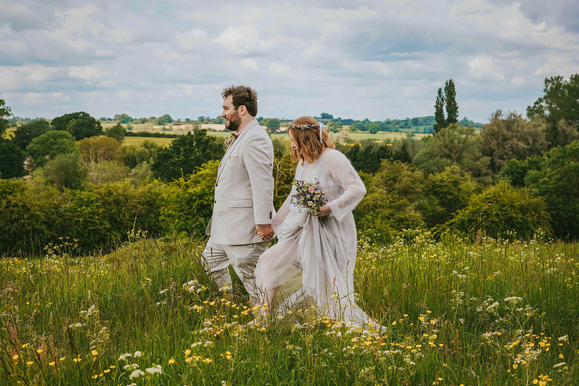 boho bride and groom wearing off white suit and dress walk through beautiful green meadow in somerton uk taken by david lefebvre a buckinghamshire wedding photographer