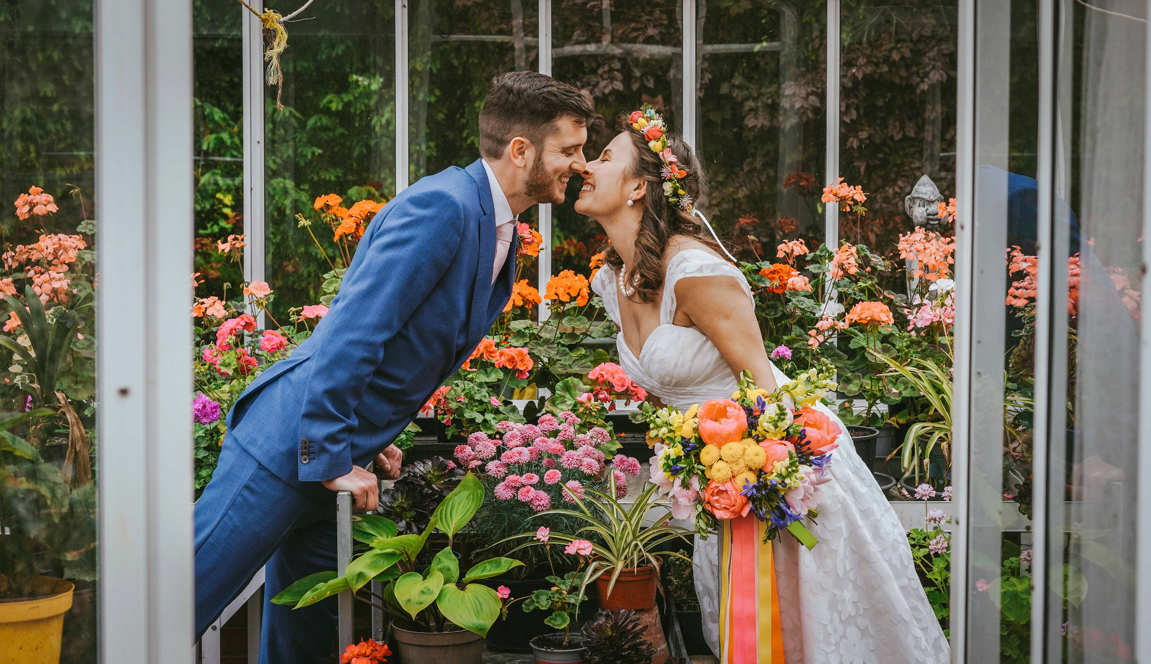 stephen the groom leans over flowers in green house about to kiss his bride gaydeliya in white with colourful vibrant bouquet and headband taken by wedding photographer in buckinghamshire david lefebvre