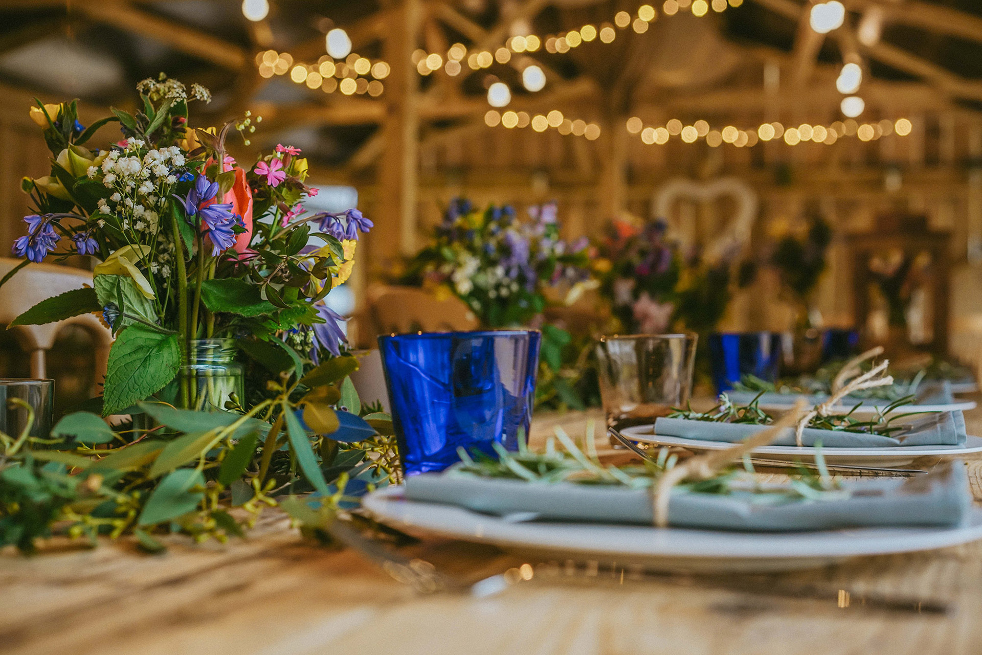 close up photo of blue violet white pink and yellow wild flowers in jars at wedding table with fairy lights and plates in the background