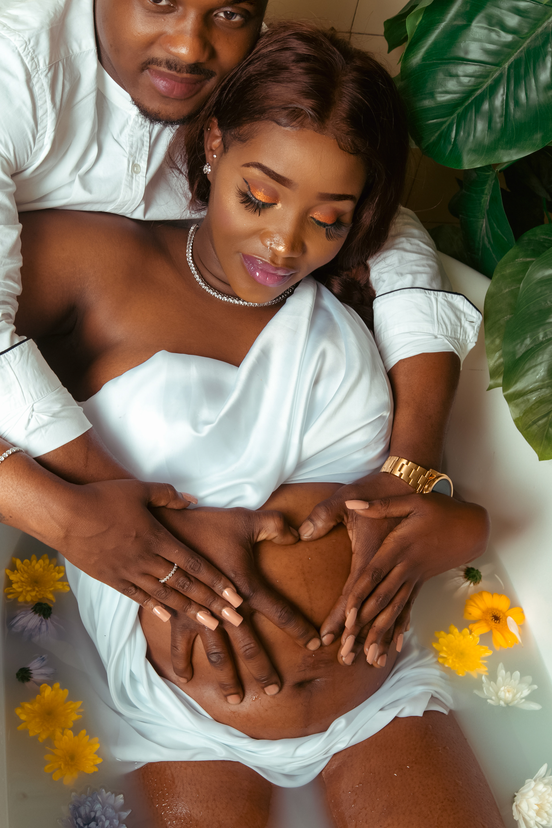 Maternity bathtub shootfeaturing the father of the baby and the mother to be holding the bump together as dad makes a heart shape gesture with his hands 