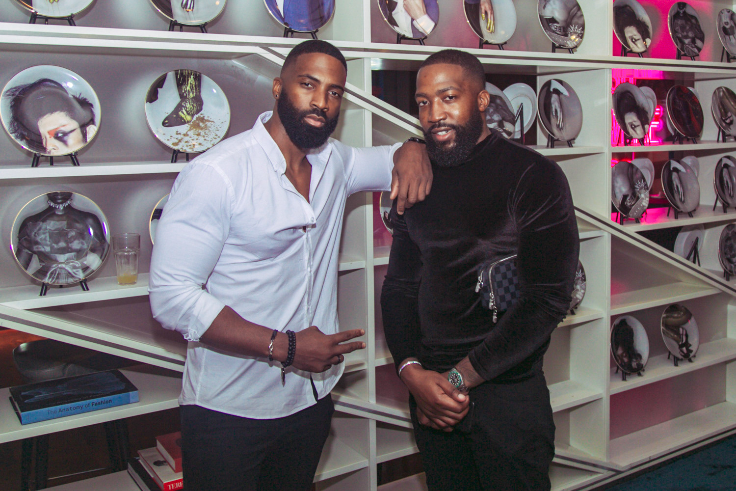 Two friends posing together infront of a wall or shelf of plates with designs and faces printed on them 