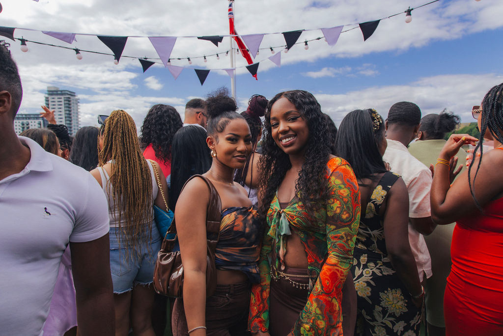 Two friends at a boat party with crowd in the background and blue skies and sun above