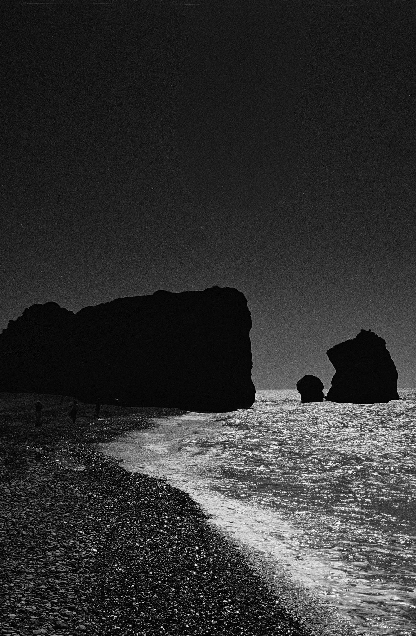 Dark sea stacks rise from a glittering shoreline as figures wander the edge of the Mediterranean in fading light.