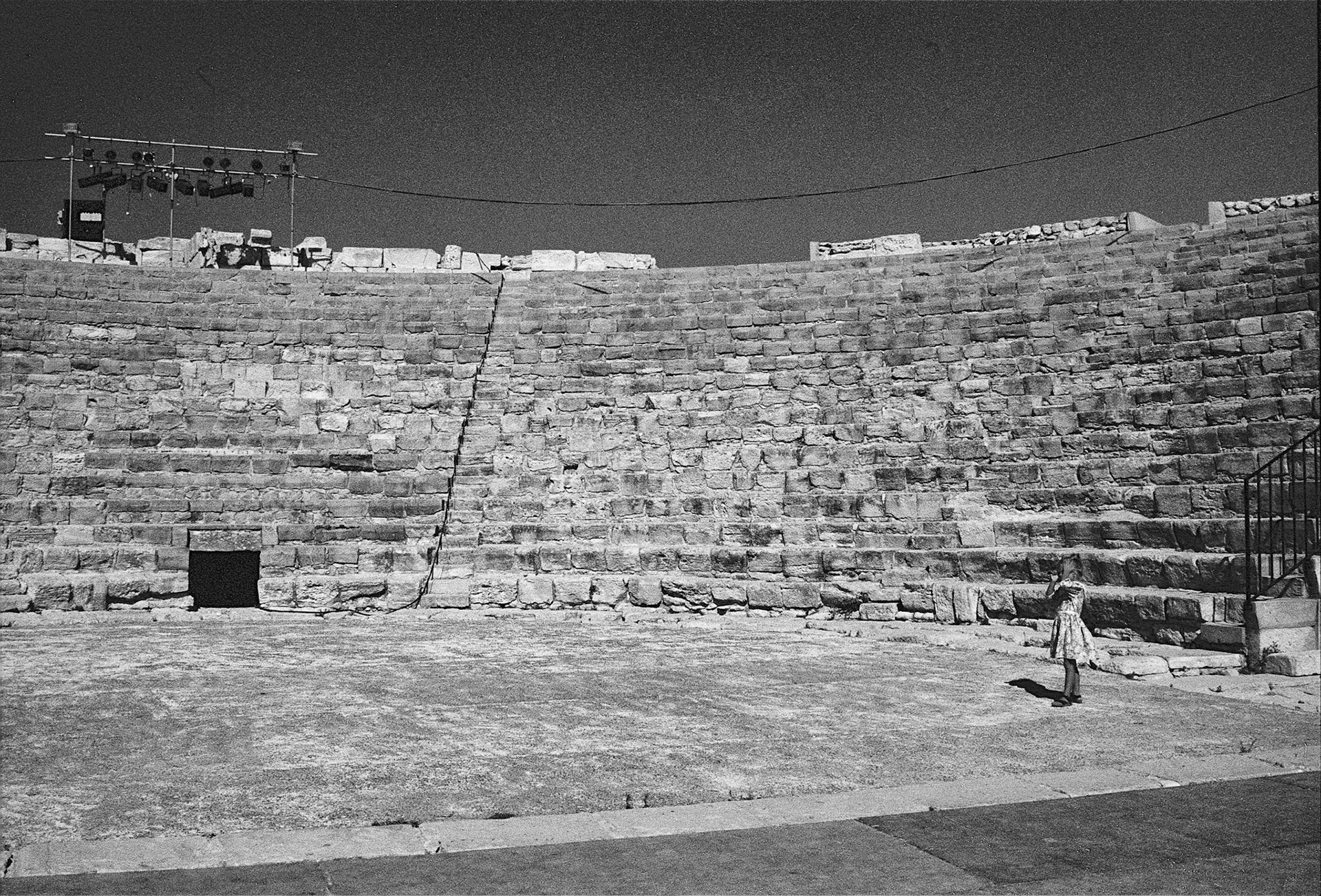 A lone figure crosses the worn stage of an ancient theatre, dwarfed by sweeping tiers of sunlit stone.
