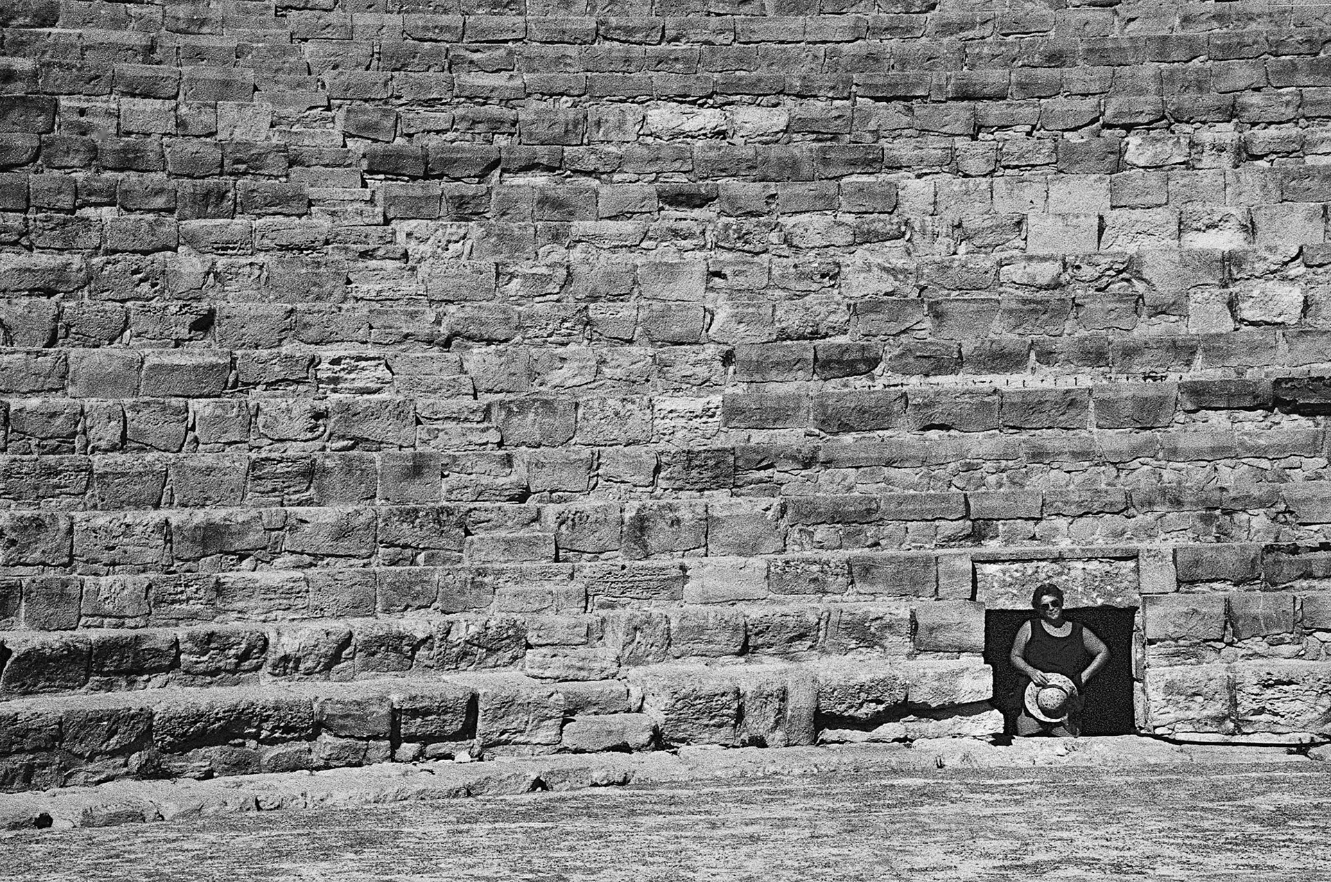 A solitary figure emerges from a small opening in an ancient stone wall, dwarfed by its scale and texture.